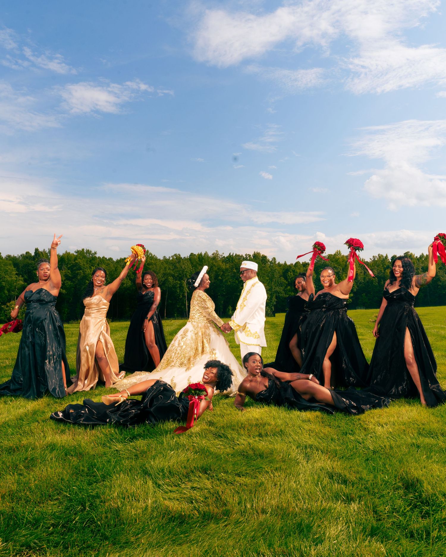 Wedding party in black gowns and white suit celebrate on green field, posing dramatically under blue sky.