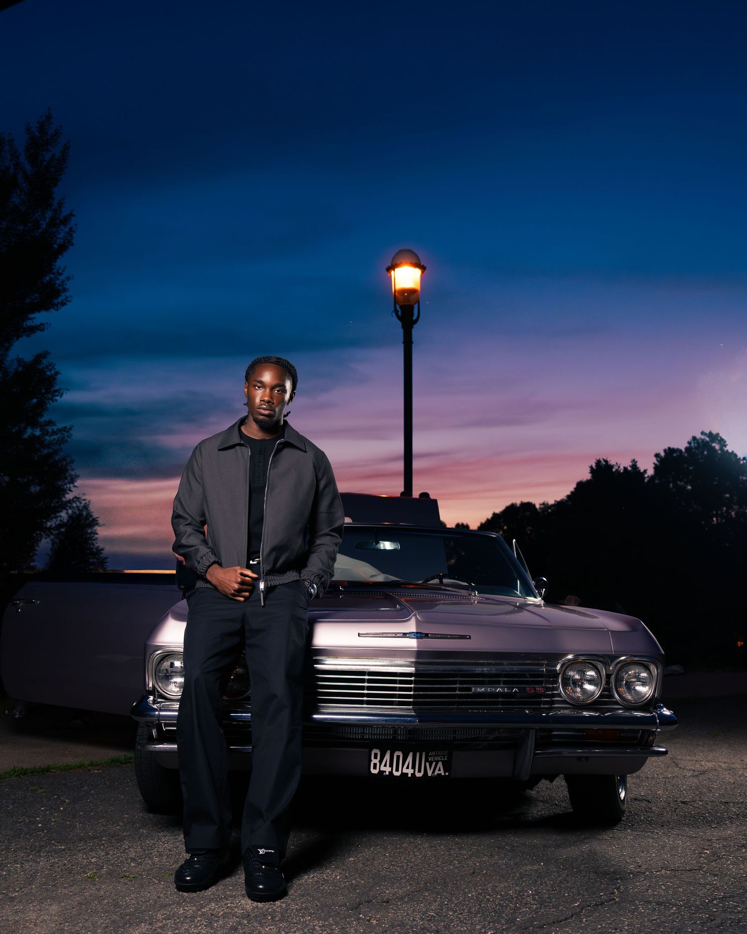 Man leaning against a classic vintage car under a street lamp at dusk with a colorful sunset sky.