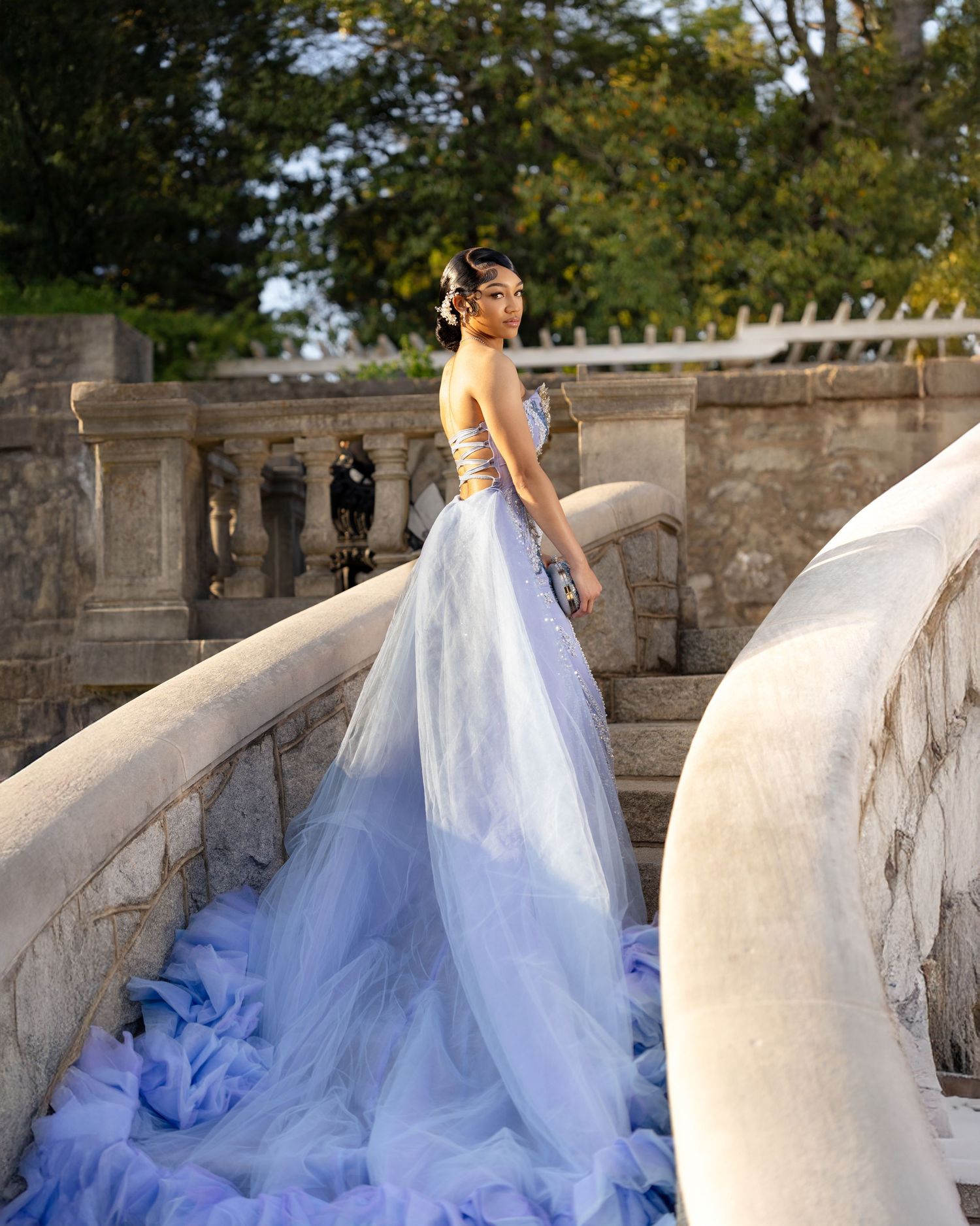 Woman in lavender tulle ball gown poses gracefully on stone staircase balustrade with lush green trees backdrop.