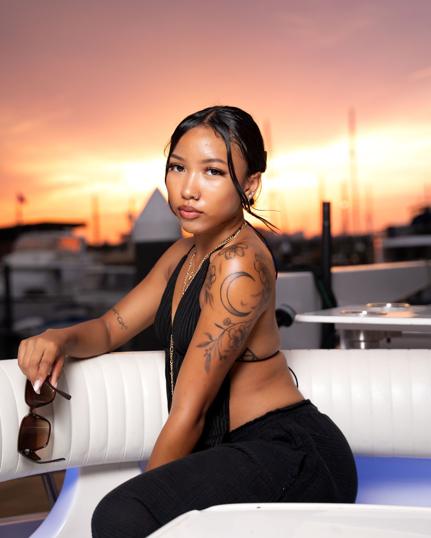 Young woman with tattoos poses on white leather sofa at rooftop venue during golden hour sunset.