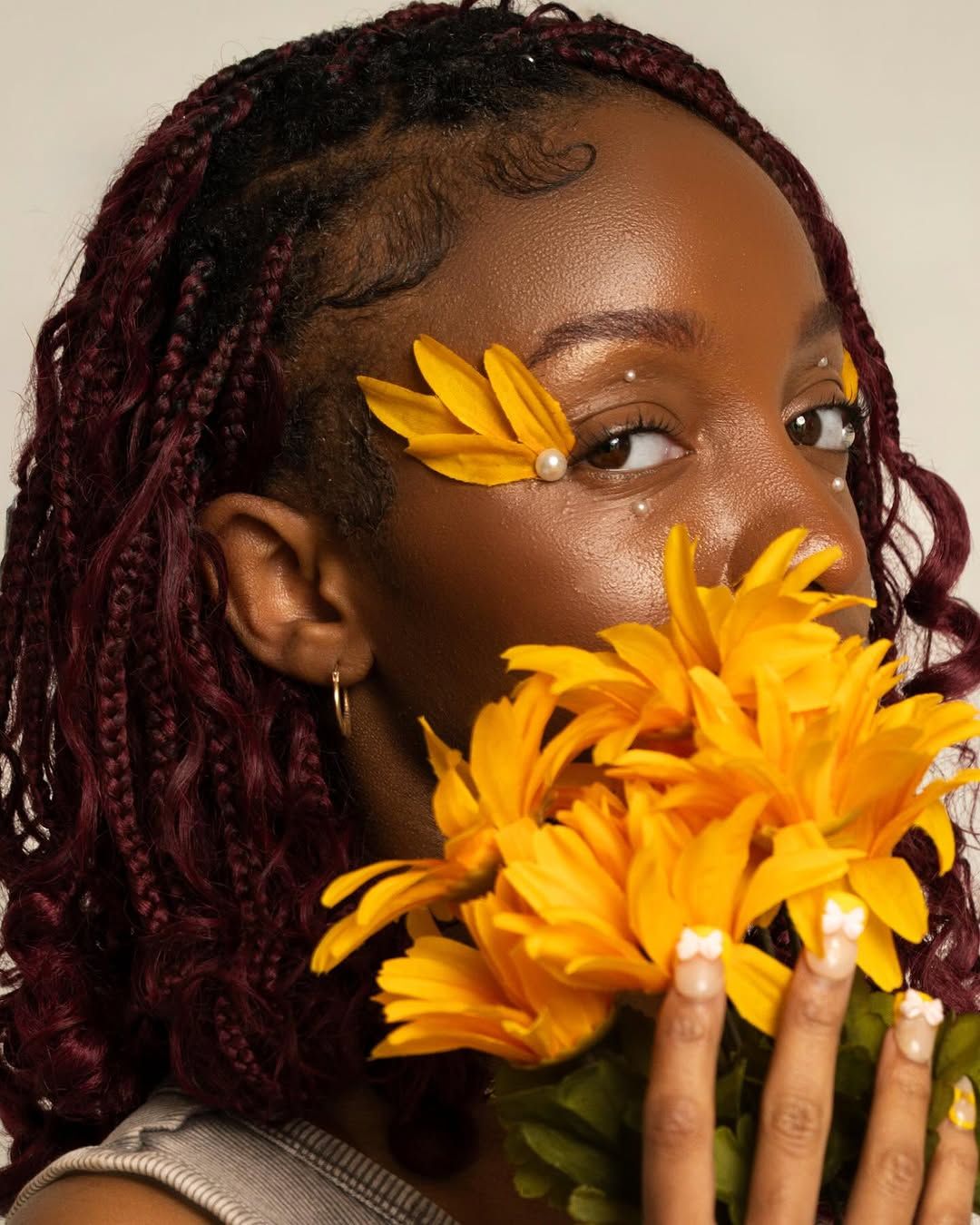 Young Black woman with burgundy braids holds yellow sunflowers, adorned with floral face gems and pearl nails.