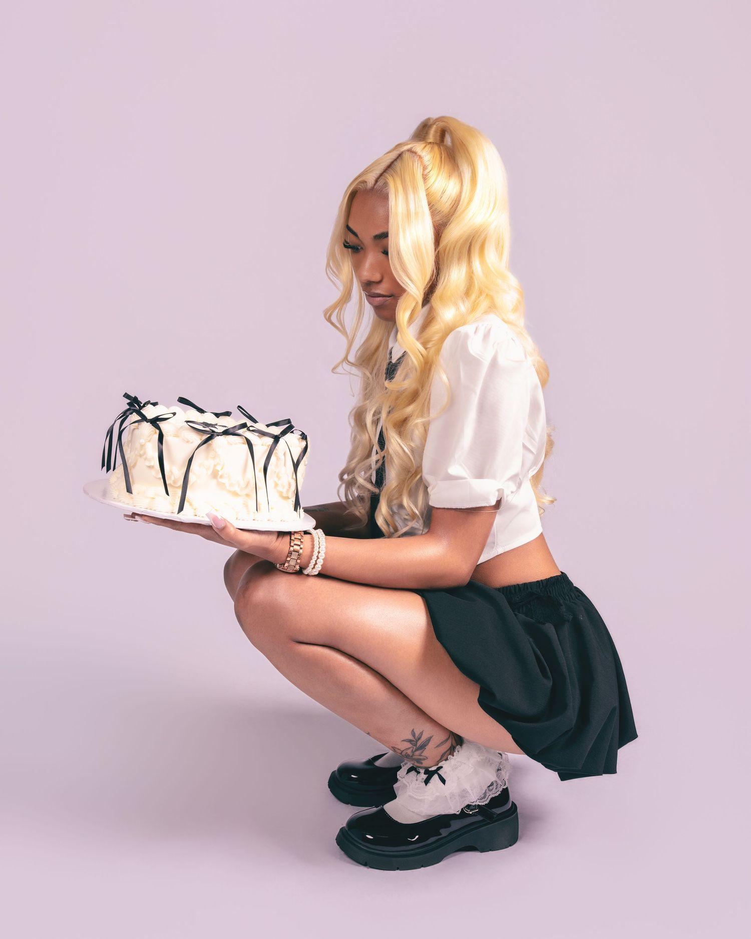 Blonde woman in white crop top and green skirt crouching while holding a white cake with chocolate drizzle.