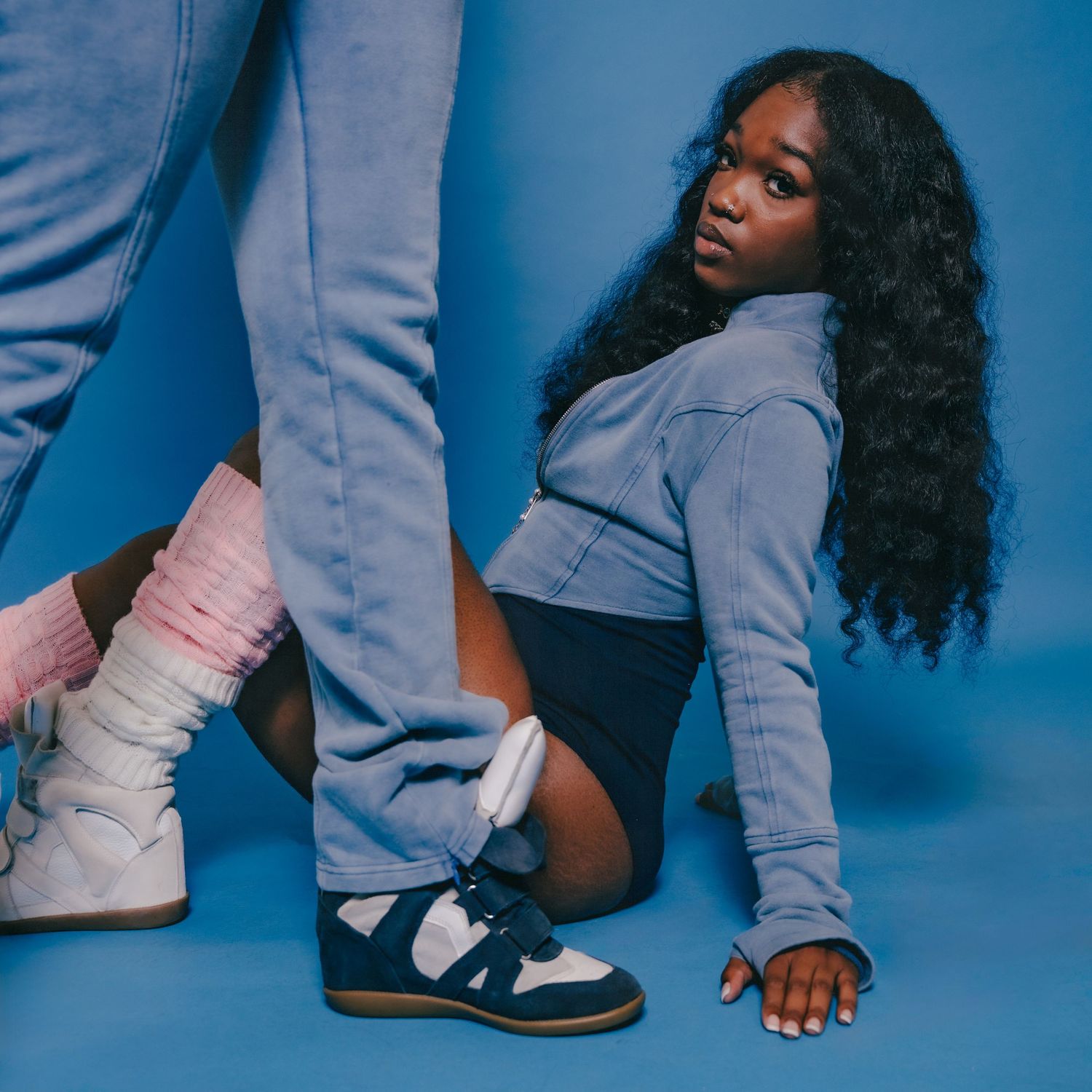 Young woman in blue jacket and dark shorts sits on floor wearing navy sneakers, styled against a blue background.