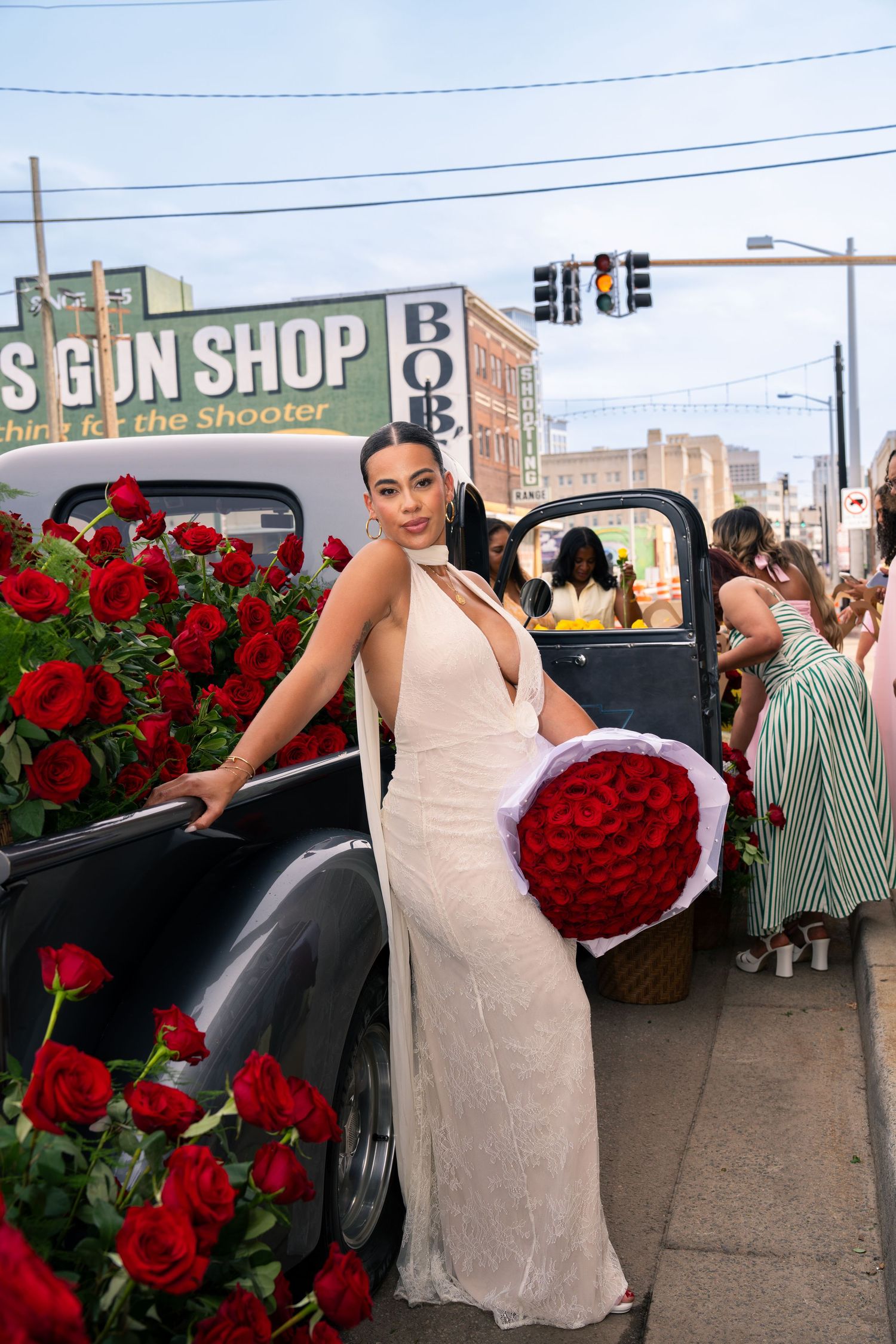 Woman in white gown holds red roses bouquet beside vintage black car decorated with roses near a gun shop.
