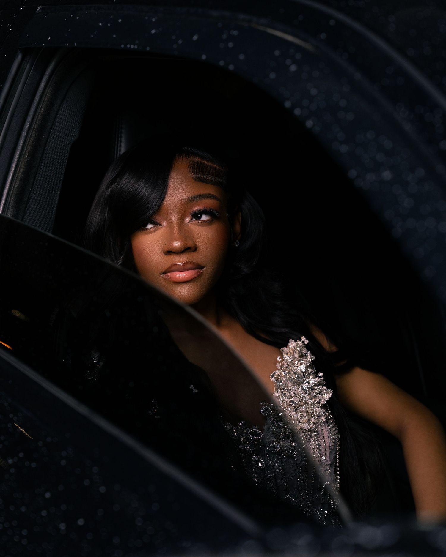 Young woman in a sparkling beaded gown gazes elegantly from a car window in a dramatic dark-toned portrait.