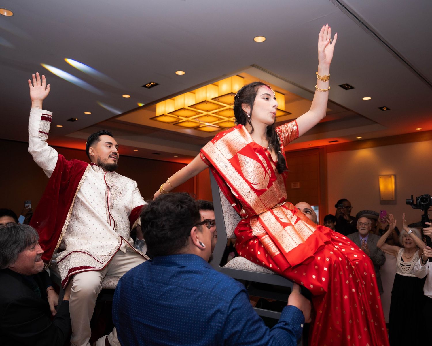 Indian couple in traditional attire lifted by guests during a joyful wedding reception dance celebration.