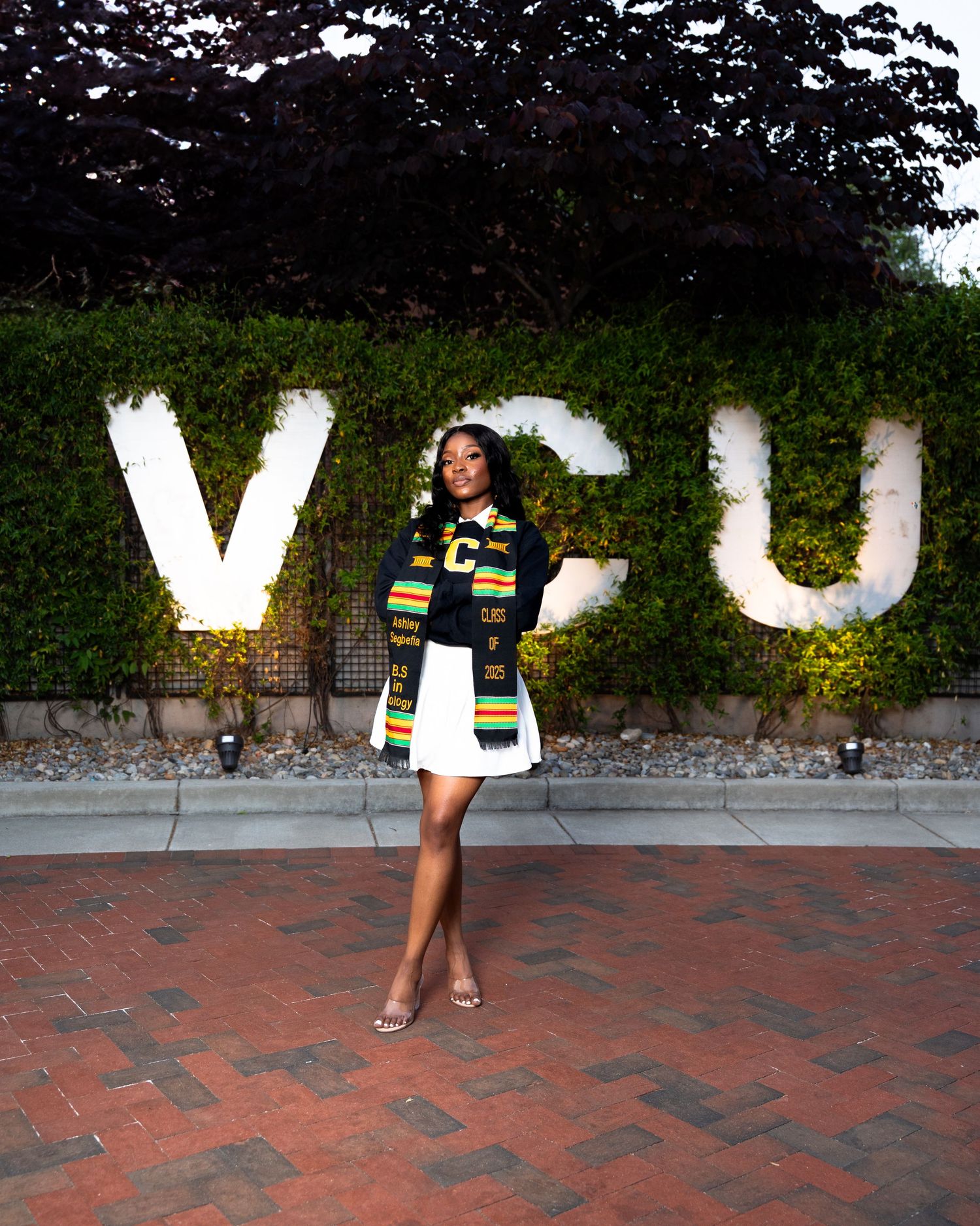 A graduate poses in a white dress and kente cloth stole in front of a VCU letter sign surrounded by greenery.