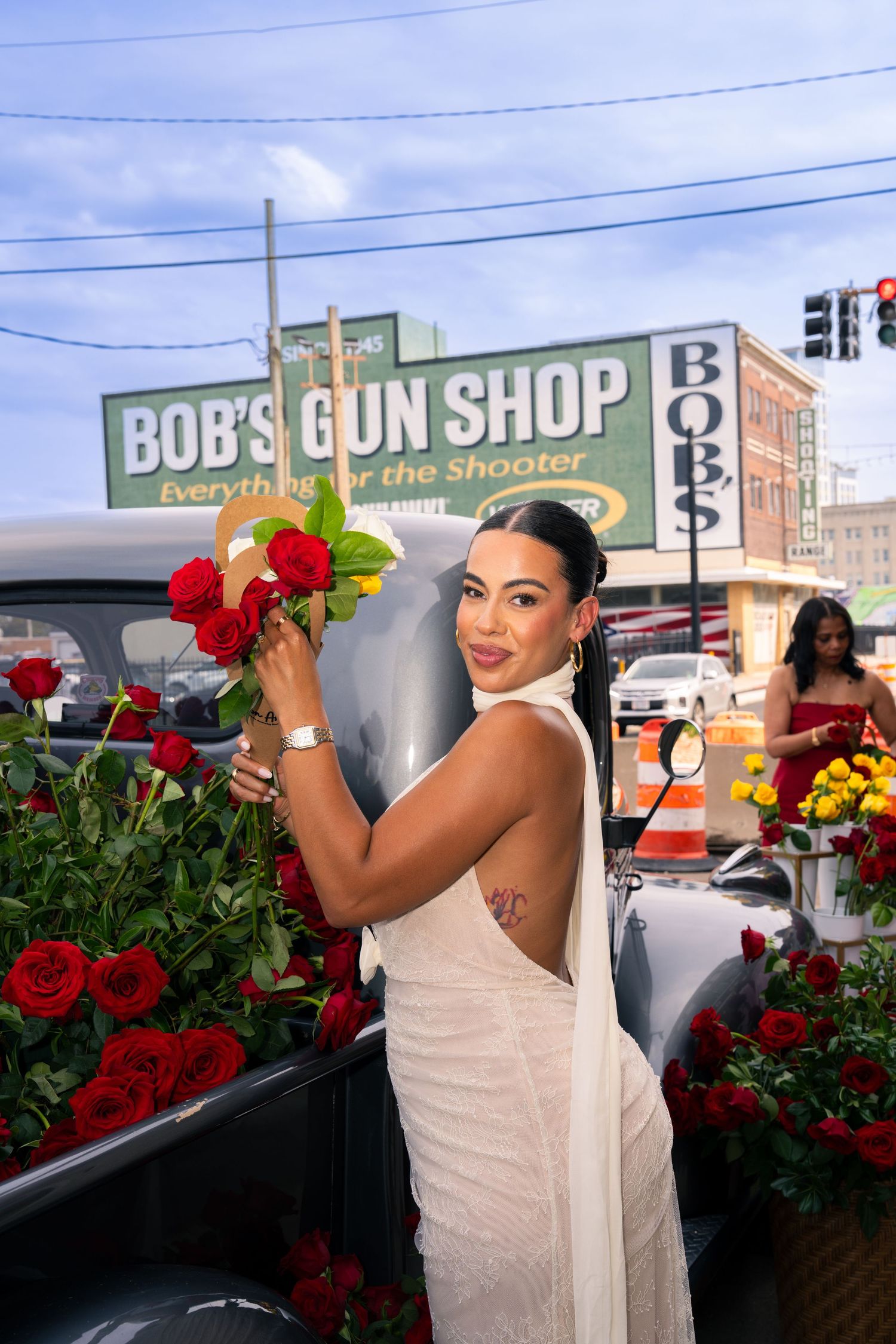 Woman in white dress holding red roses near Bob's Gun Shop sign with flowers in truck bed.