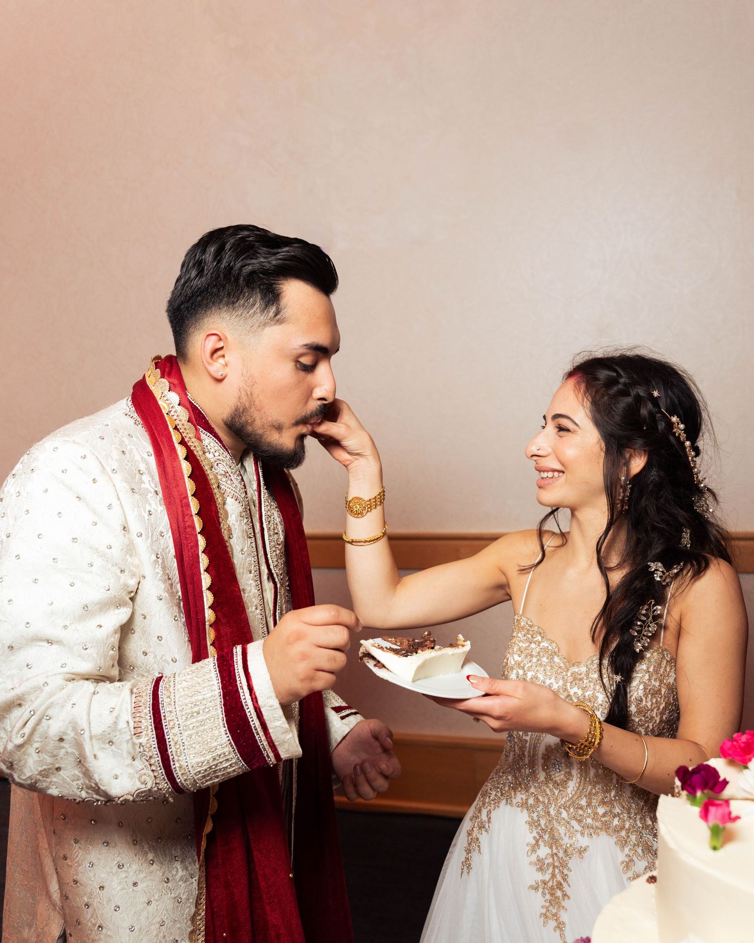 Bride in gold lehenga feeds wedding cake to groom in white sherwani with red dupatta during Indian fusion wedding ceremony.