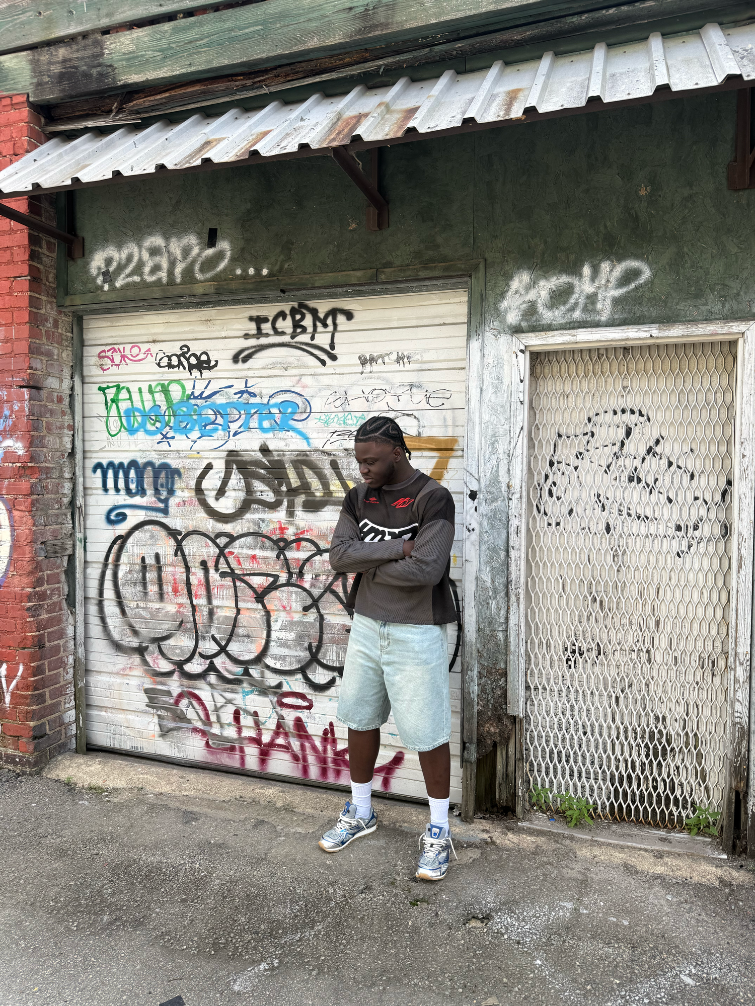 Young man in brown hoodie and shorts poses arms crossed in front of graffiti-covered garage door in urban alley.