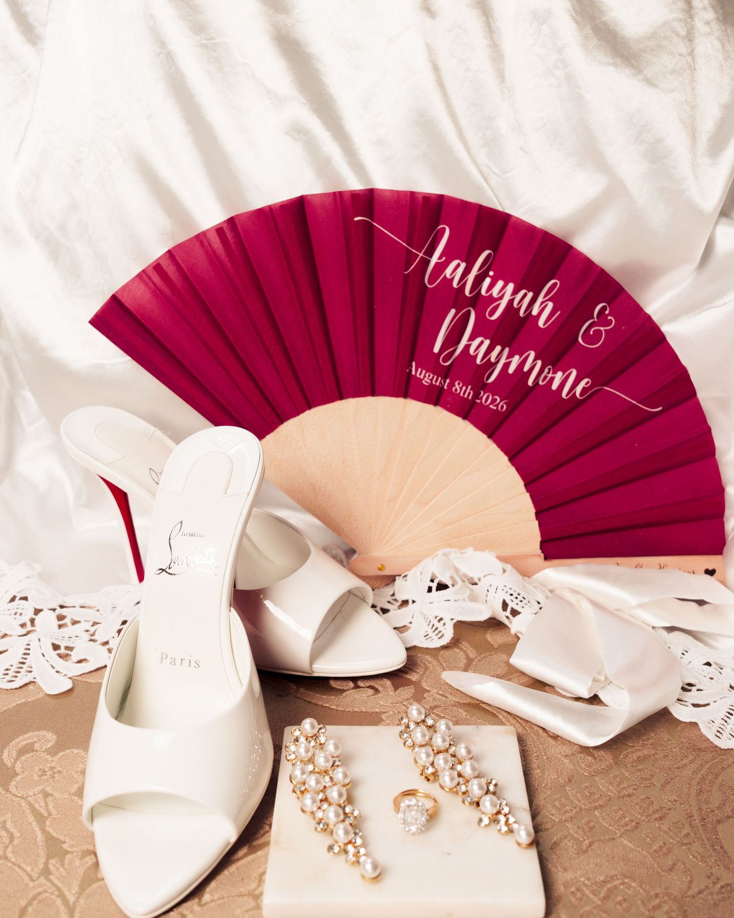Elegant wedding flat lay with red fan reading Aaliyah & Daymone, white heels, pearl earrings, and lace details.