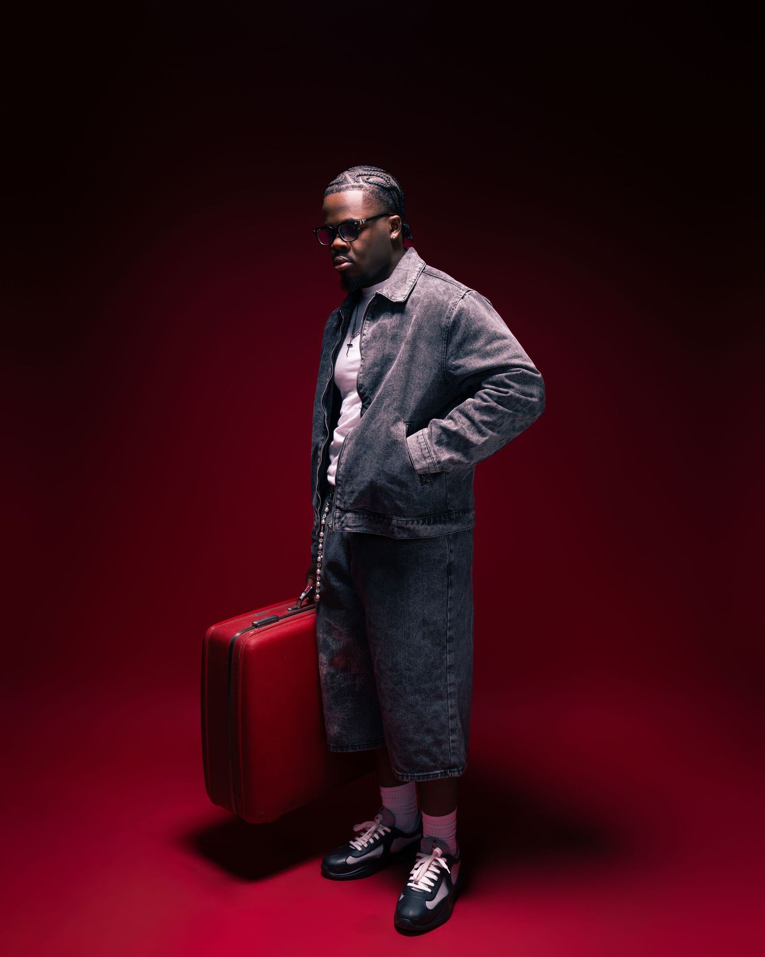 Person in gray coat and shorts holding a red suitcase against a dark red background.