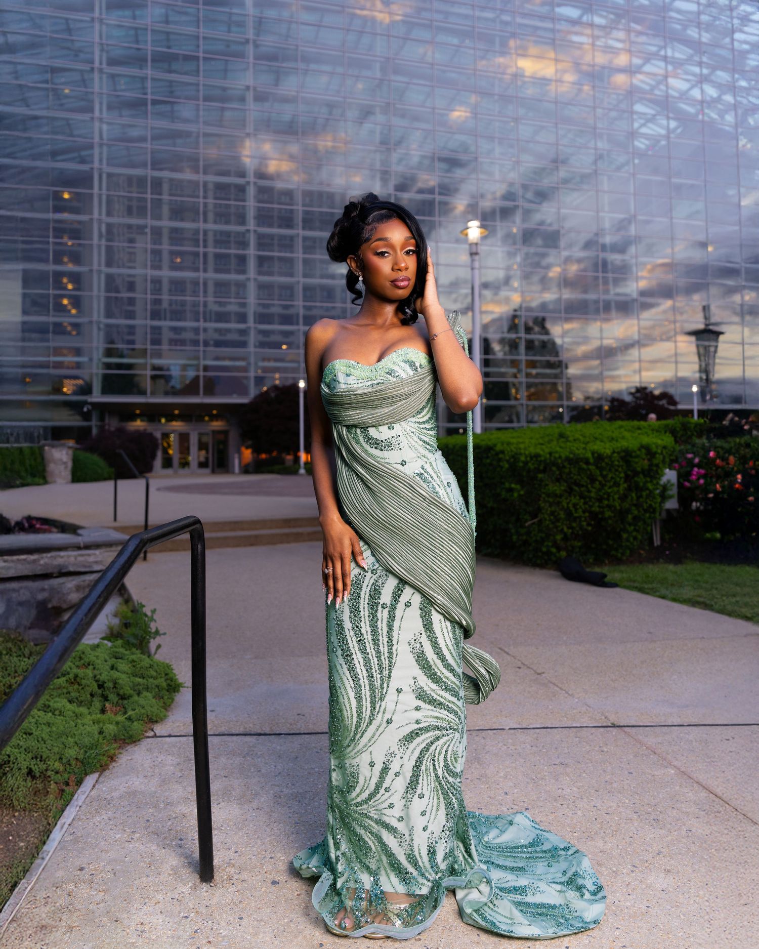 Woman in elegant sage green sequined strapless gown posing outdoors at dusk near a glass building.