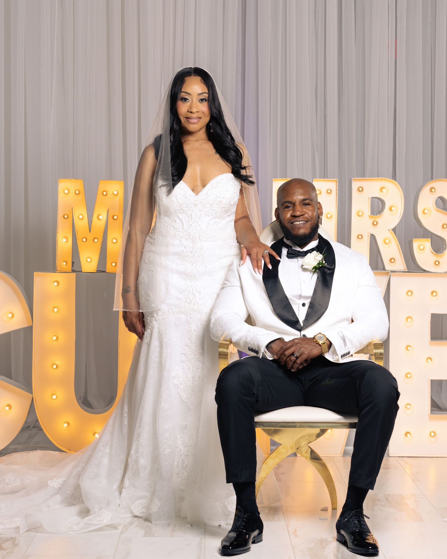 Bride in white mermaid gown stands beside groom in white tuxedo jacket at elegant wedding reception.