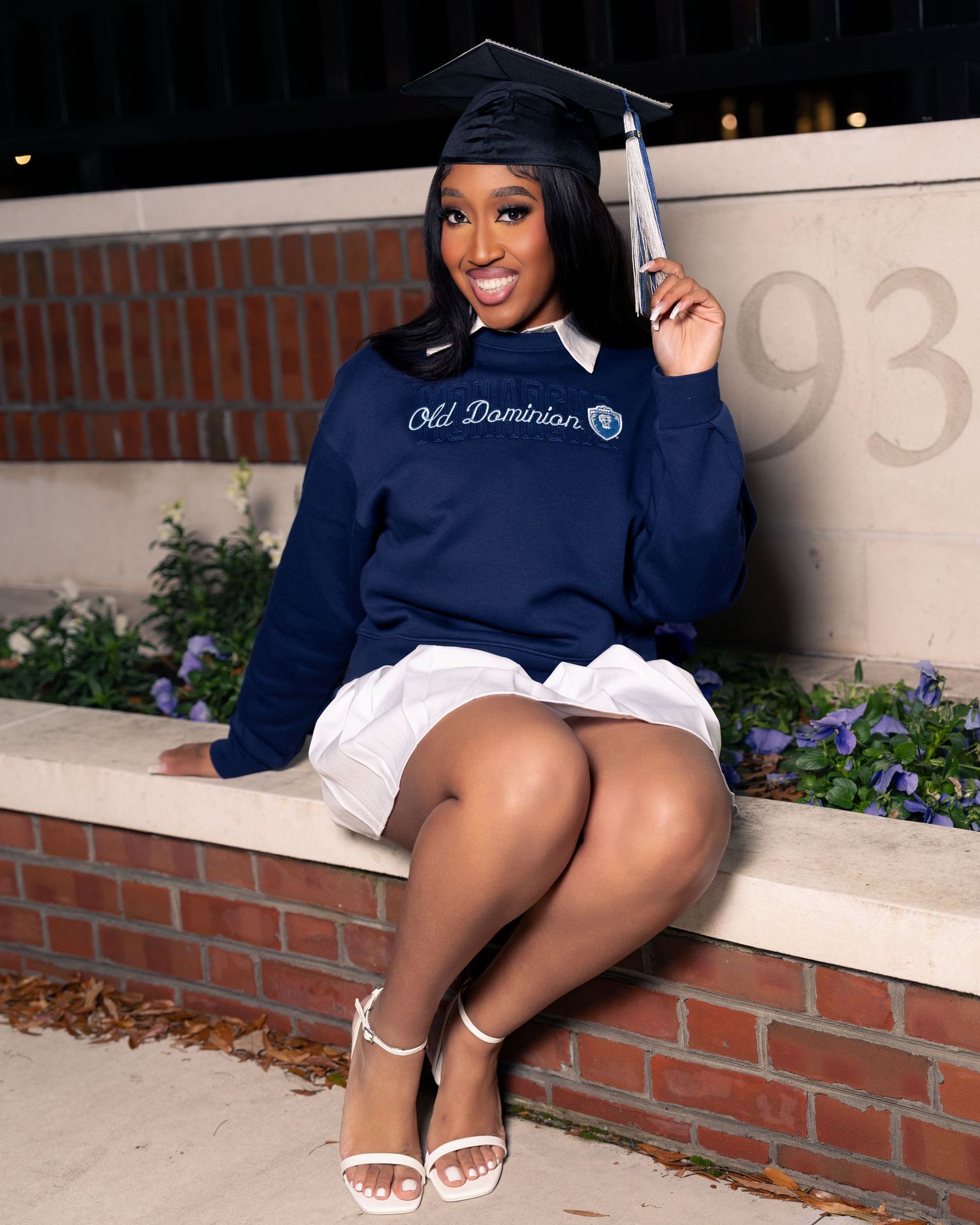 A smiling graduate in cap and Old Dominion University sweatshirt sits on a brick wall holding a diploma.