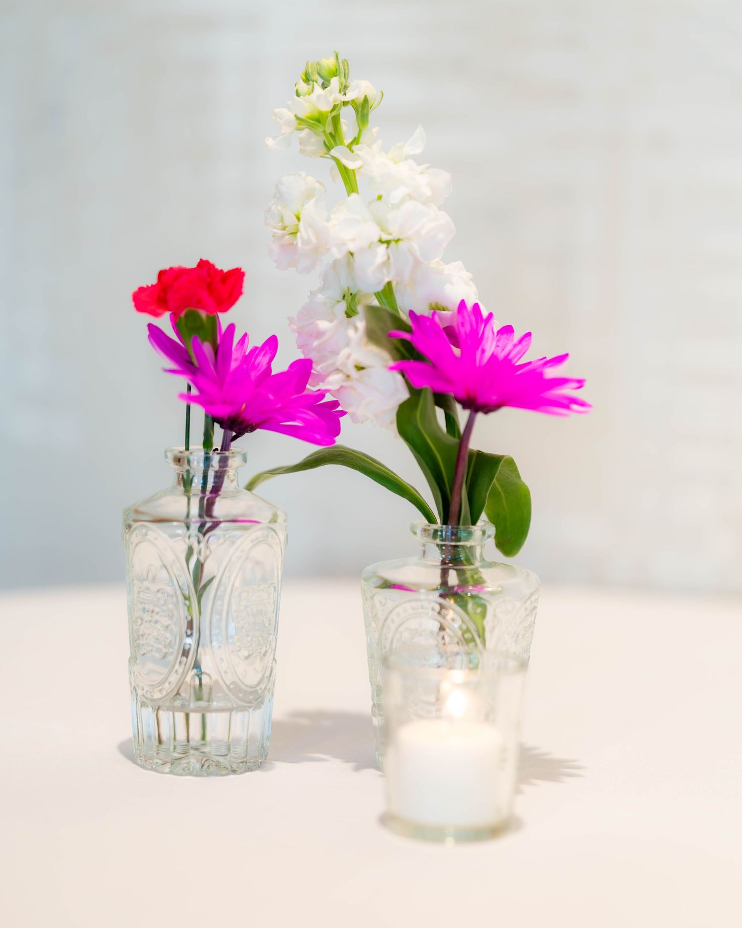 Two glass vases with pink daisy and red carnation flowers beside white blooms and a small candle on a white table.