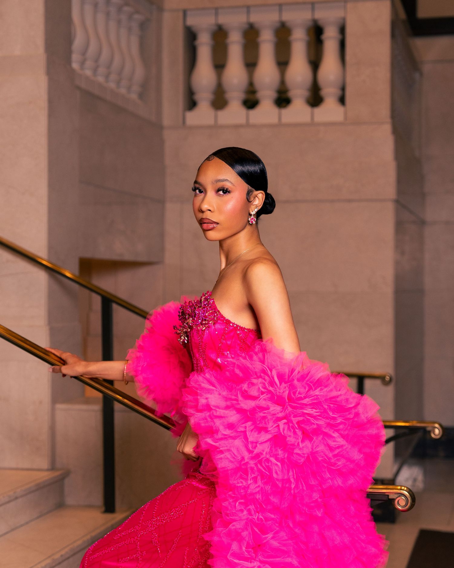 A young woman poses on marble stairs wearing a stunning hot pink ruffled tulle gown with floral embellishments.