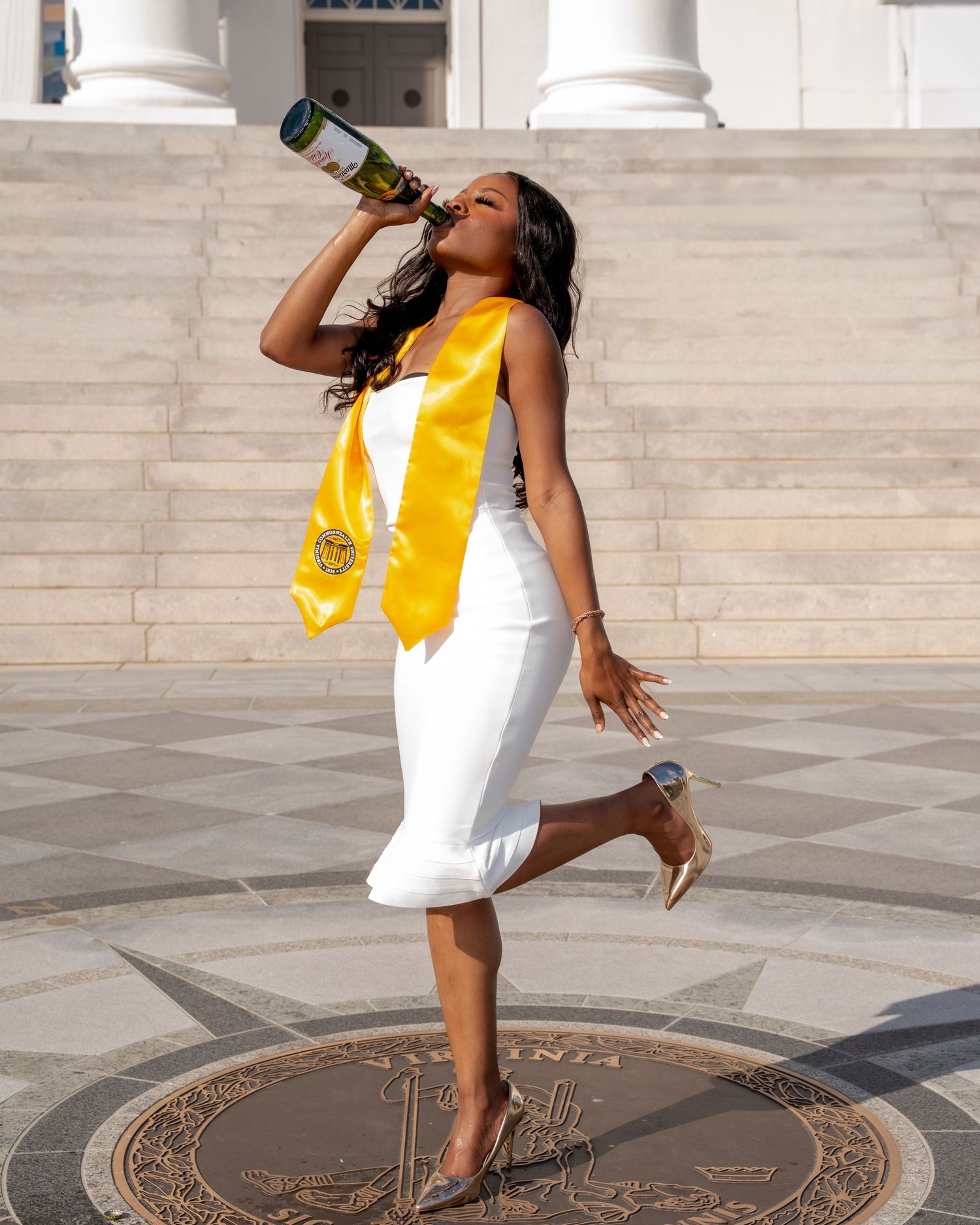 Graduate in white dress and yellow sash celebrates outdoors by drinking from a bottle on one leg.