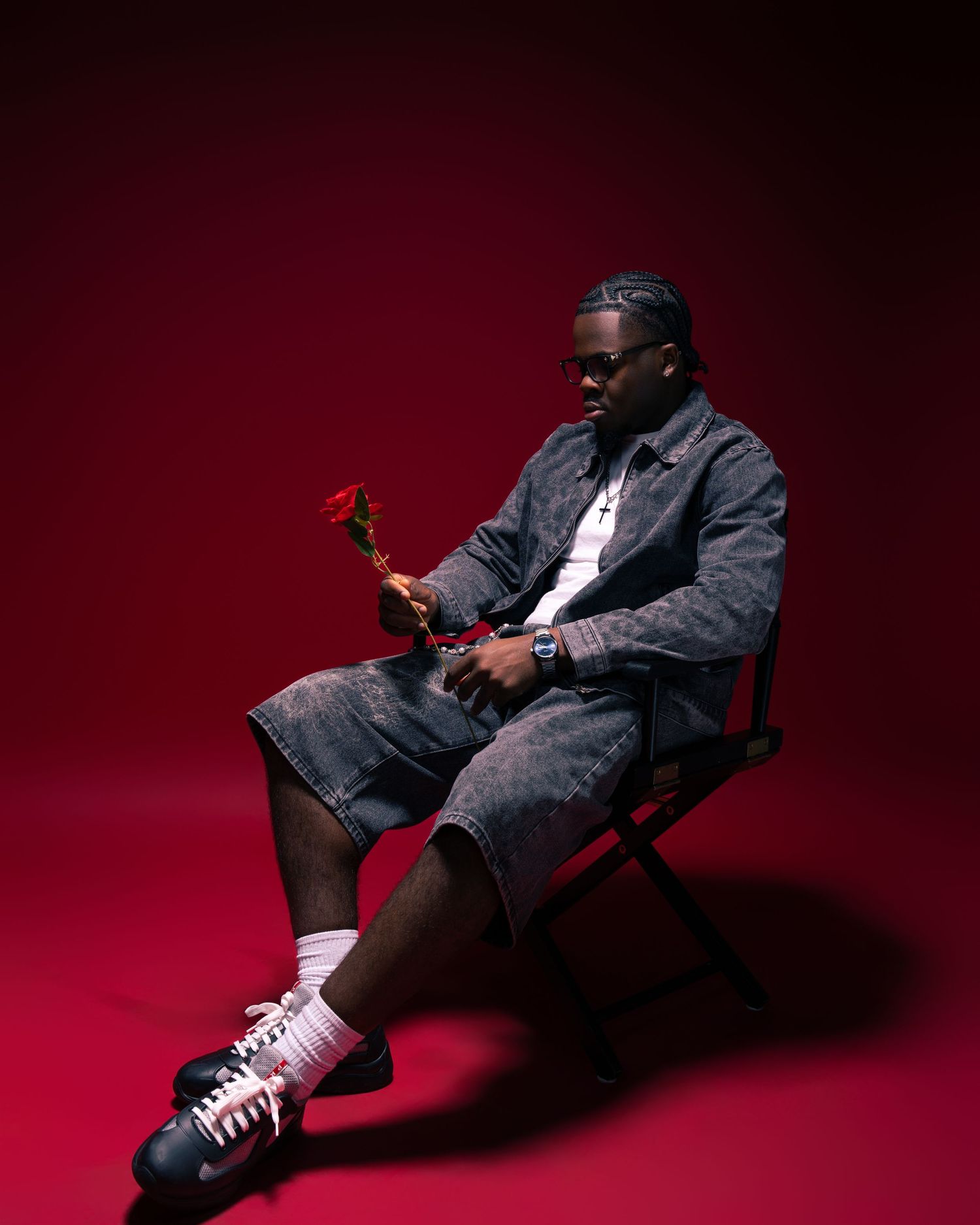 Man in denim outfit sits on chair holding red rose against vibrant red background, wearing sneakers and white socks.