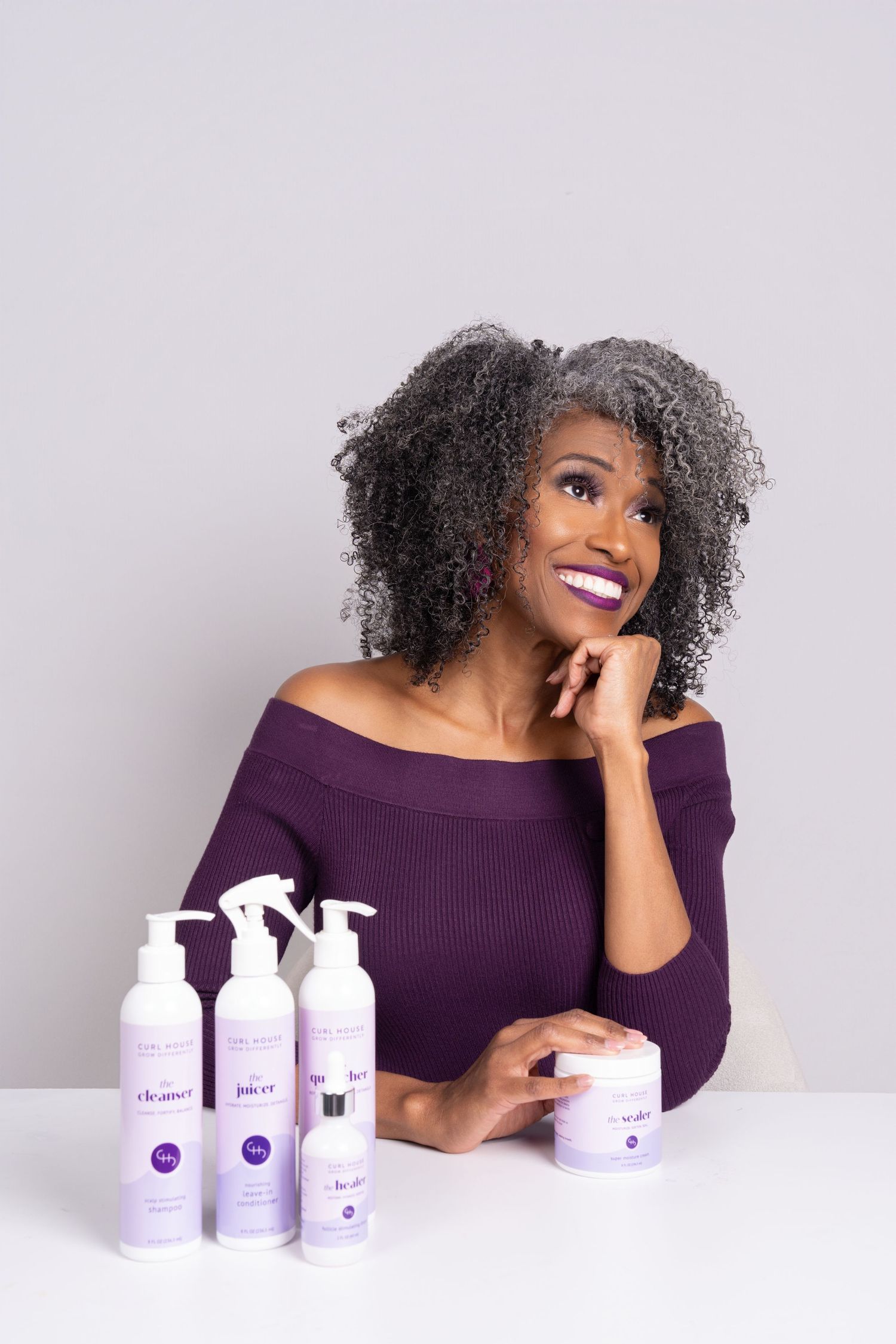 Smiling woman with natural gray curly hair in purple off-shoulder top posing with purple hair care product line.