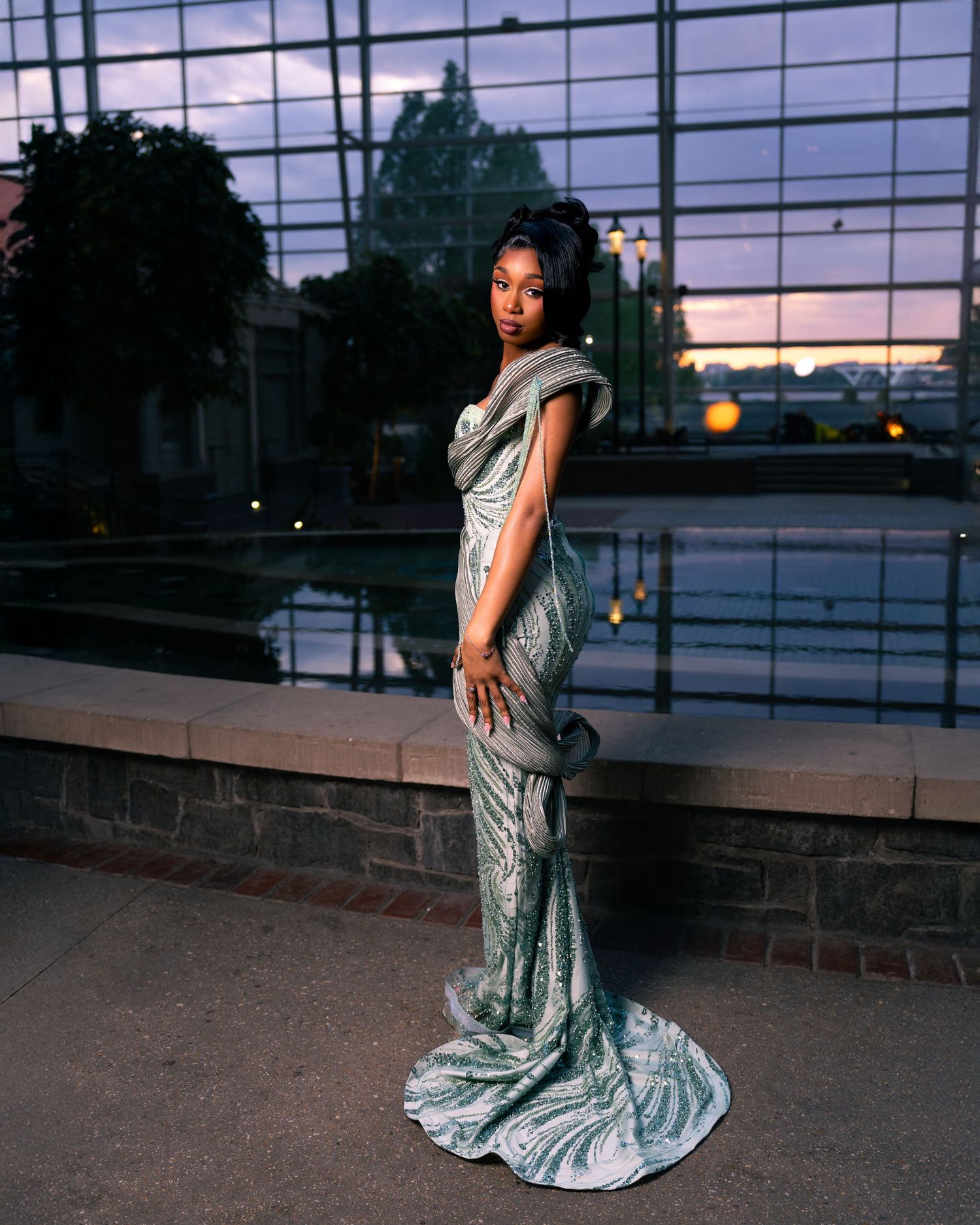 Woman in elegant silver mermaid gown poses outdoors at dusk near reflective glass building.