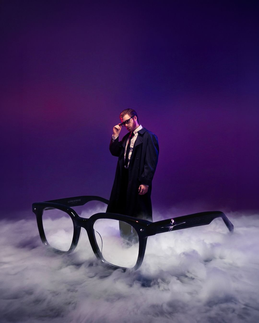 Man in black coat poses behind oversized black glasses amid purple smoke for a dramatic eyewear advertisement.