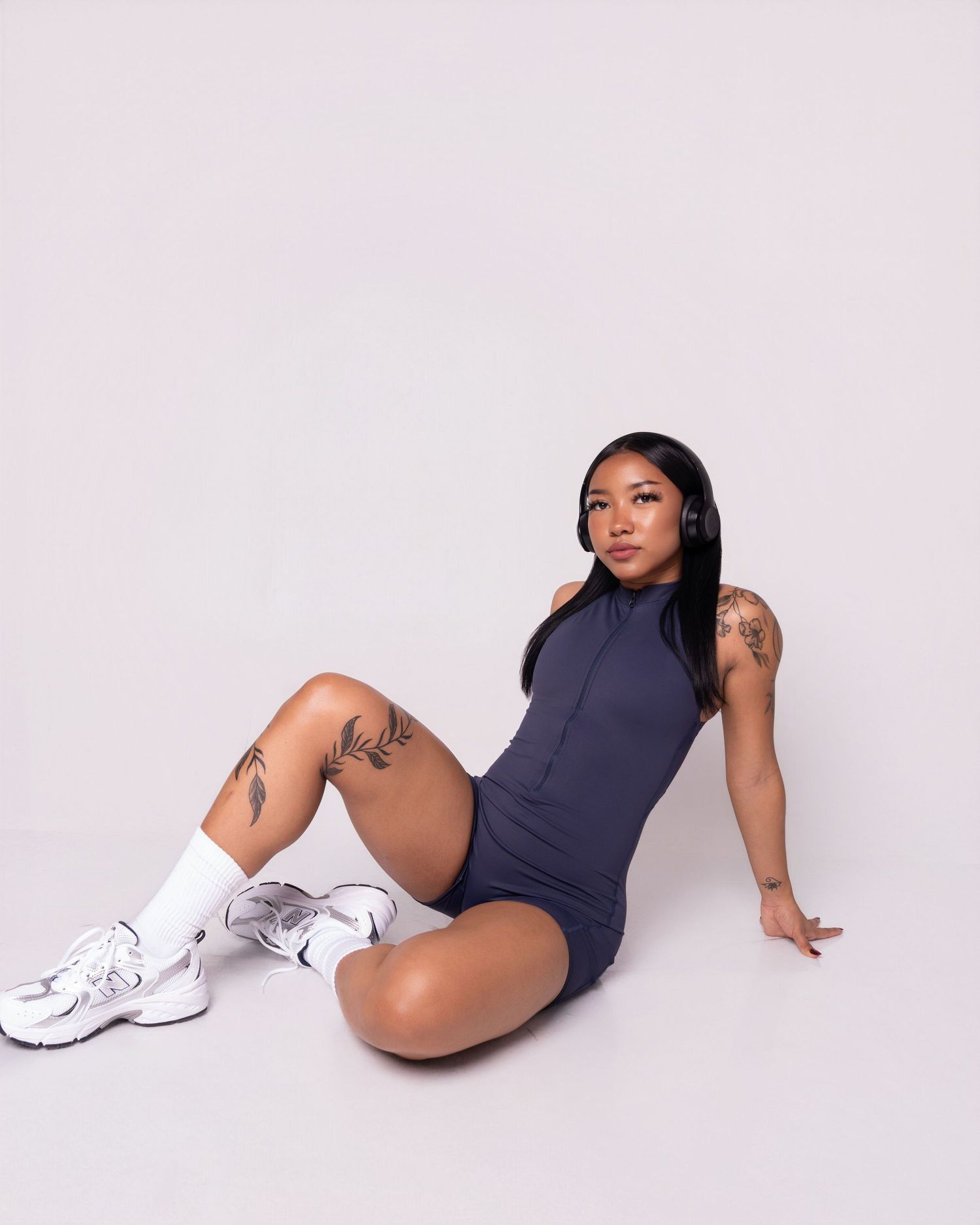 Woman with tattoos sitting on floor wearing dark bodysuit, white socks and sneakers against light background.