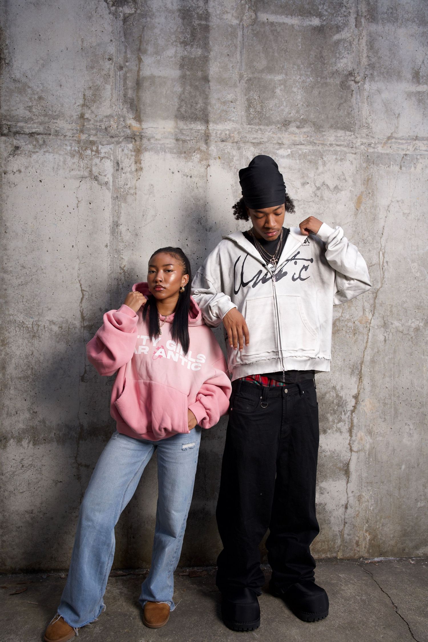 Two young people in streetwear pose against a concrete wall, wearing oversized hoodies and baggy jeans.