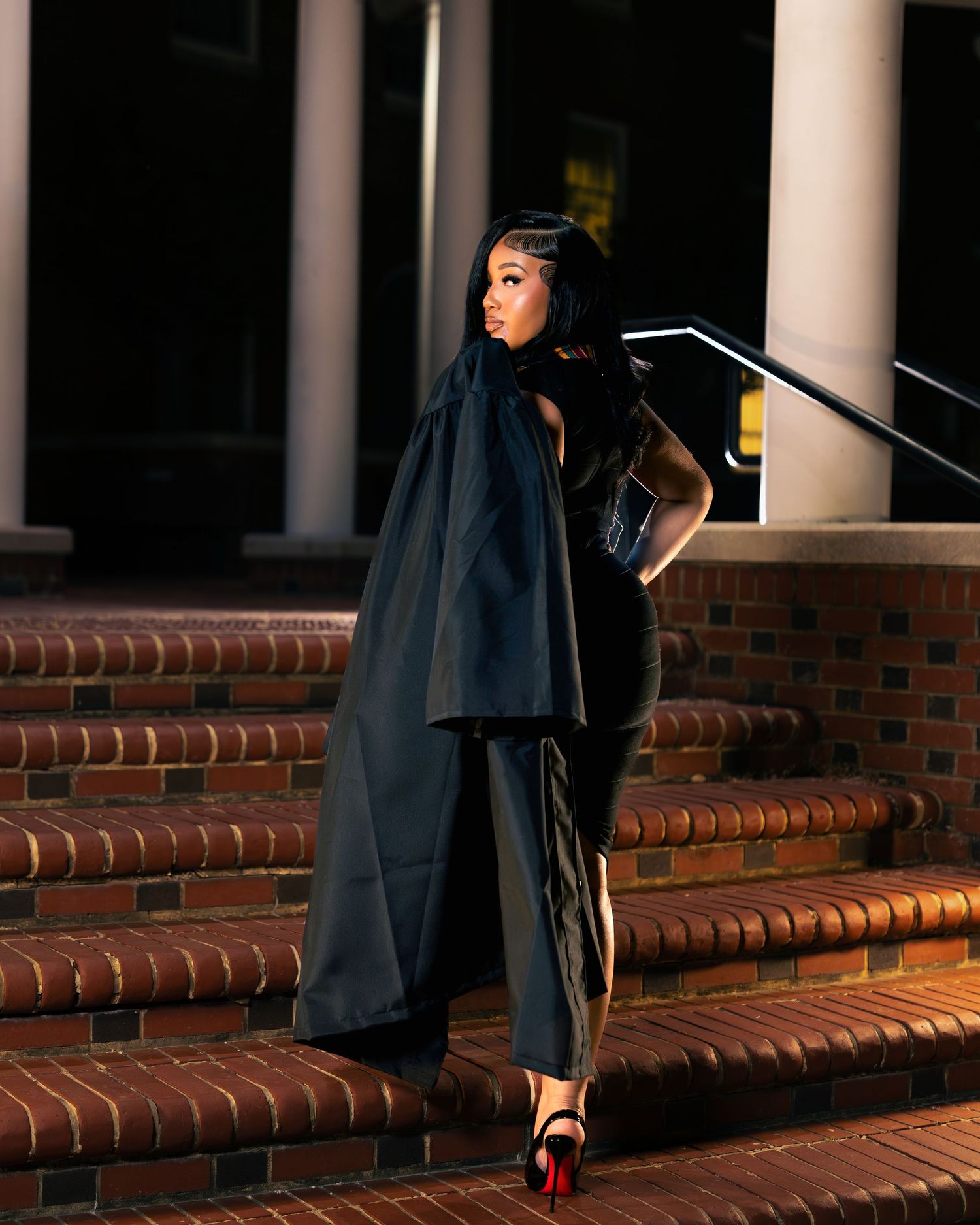 Woman in elegant black cape outfit and red heels poses on brick steps outside a building at night.
