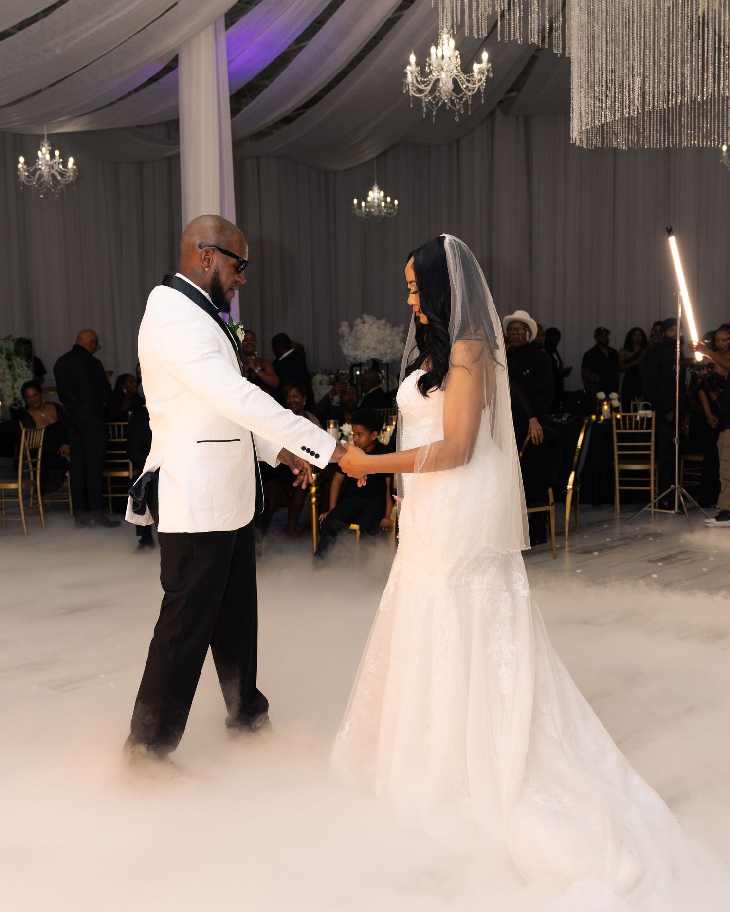 Bride and groom share first dance surrounded by fog machine clouds in elegant chandelier-lit wedding reception venue.