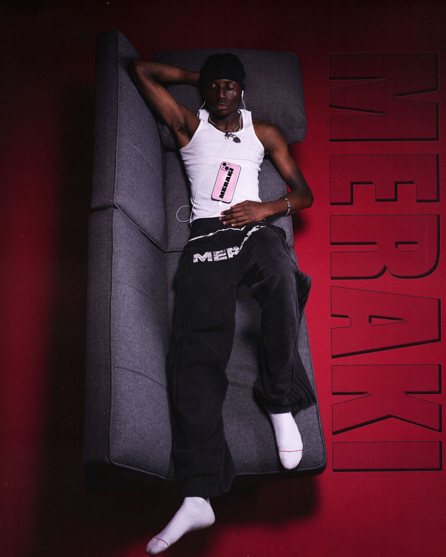 Young man in white tank top and dark Meraki sweatpants lying on gray sofa against red background.