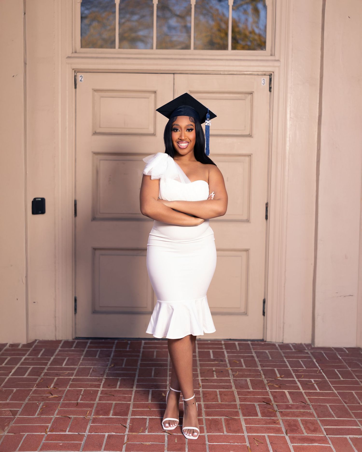 Young woman in white one-shoulder dress and graduation cap smiling in front of beige double doors on brick patio.