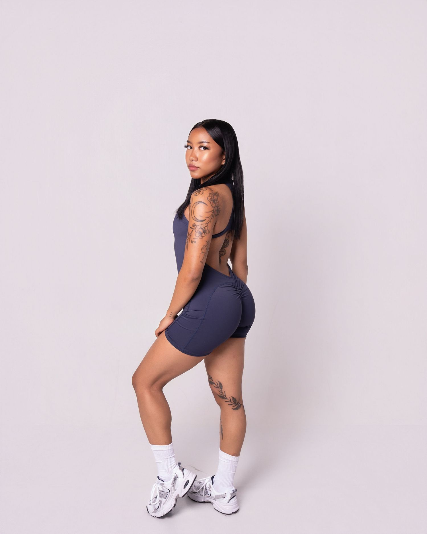 Woman in navy blue athletic shorts and top posing confidently, showing tattoos, wearing white sneakers.