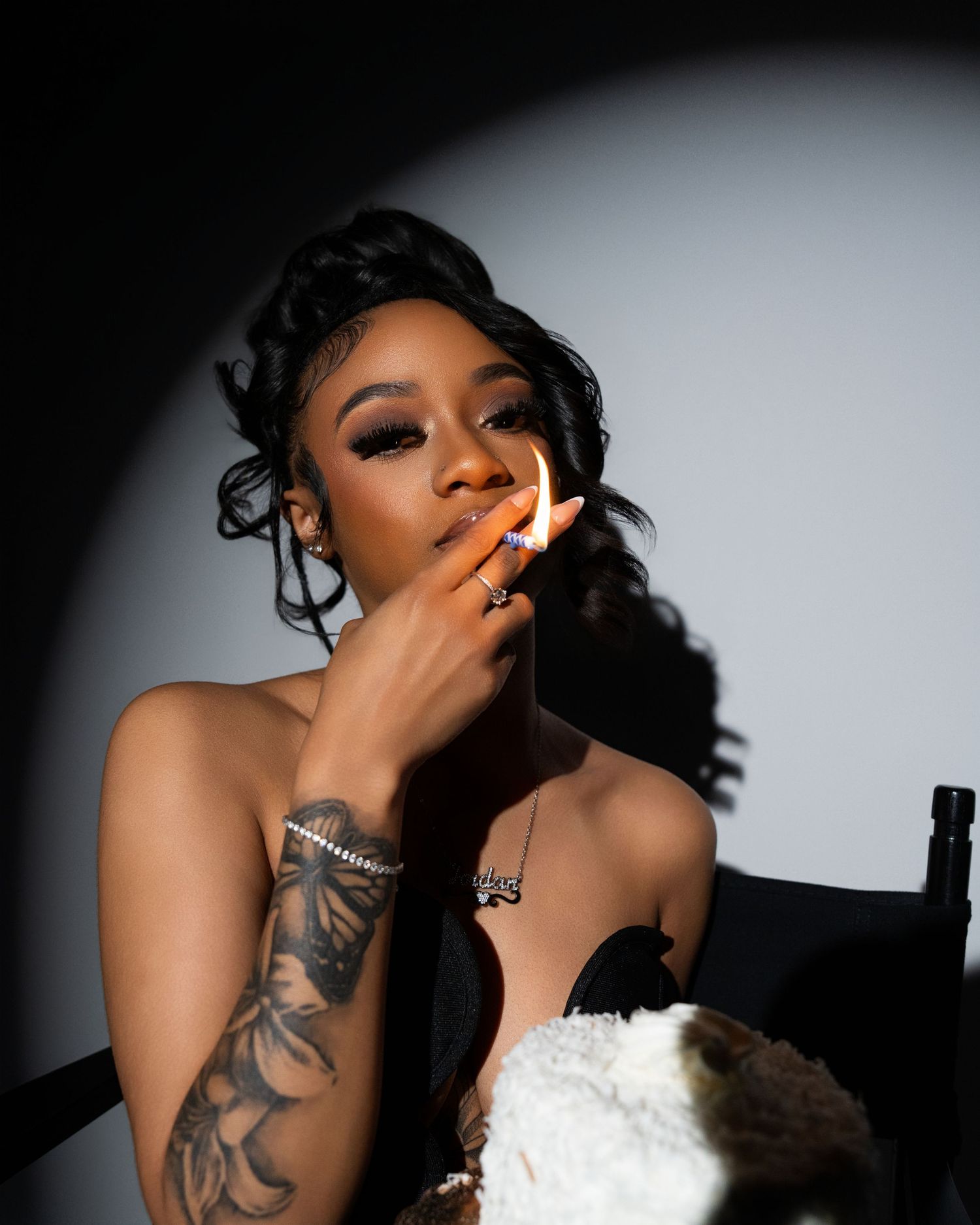 Young woman with tattoos and curly hair lighting a cigarette, wearing a strapless top in dramatic studio lighting.