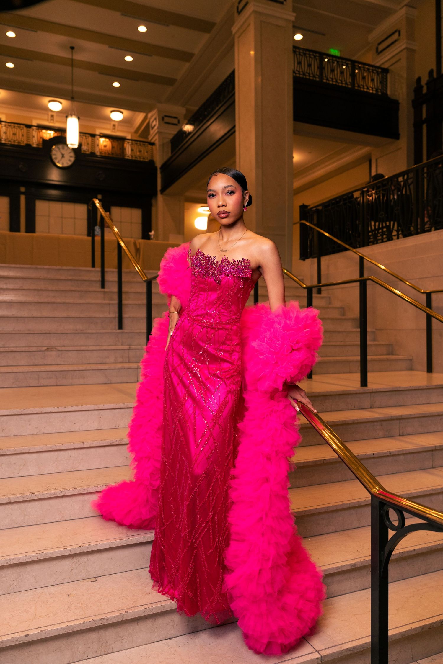 Woman in a stunning hot pink strapless beaded gown with feather trim poses elegantly on grand staircase.