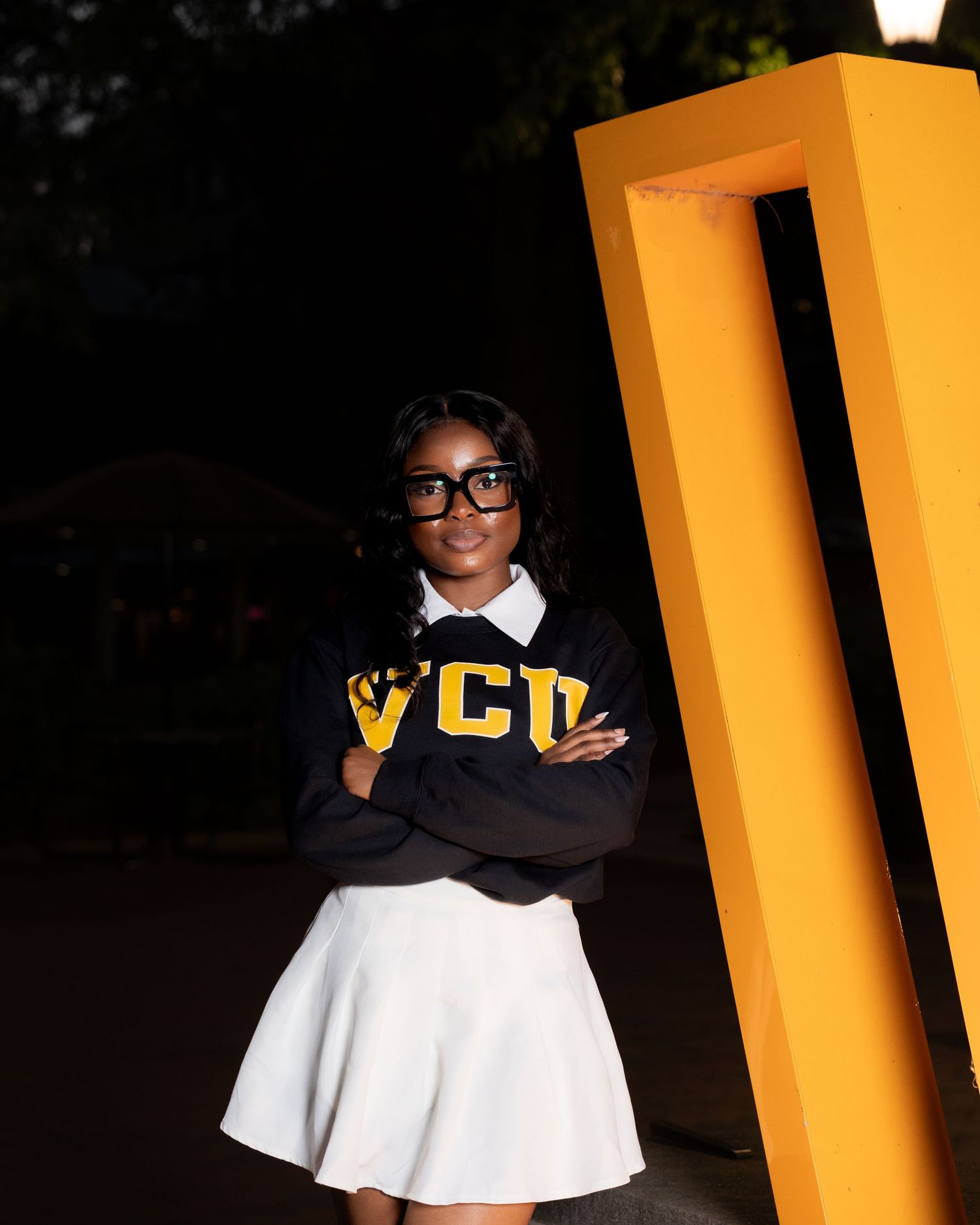 Young woman in VCU sweatshirt and white skirt poses confidently beside a yellow structure at night.