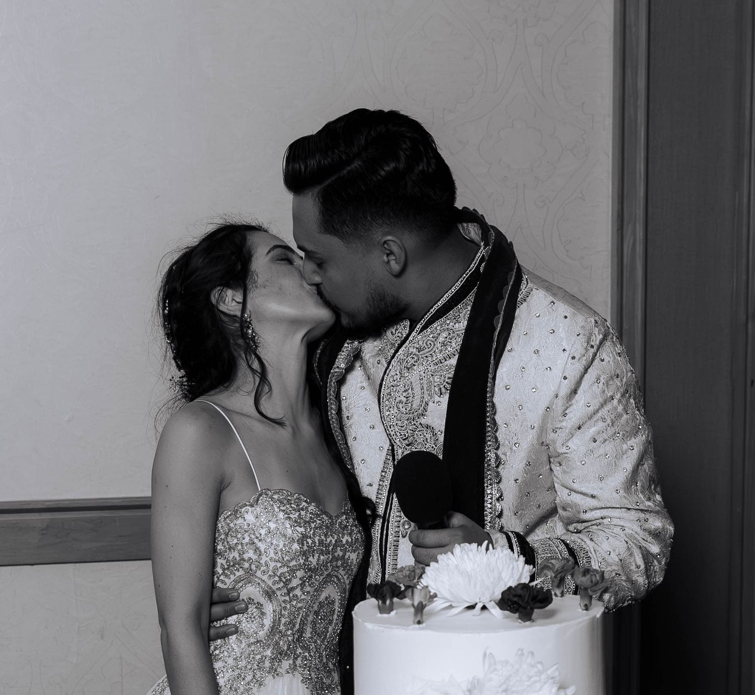 Black and white photo of a couple sharing a kiss at their wedding reception, with a white cake in front of them.