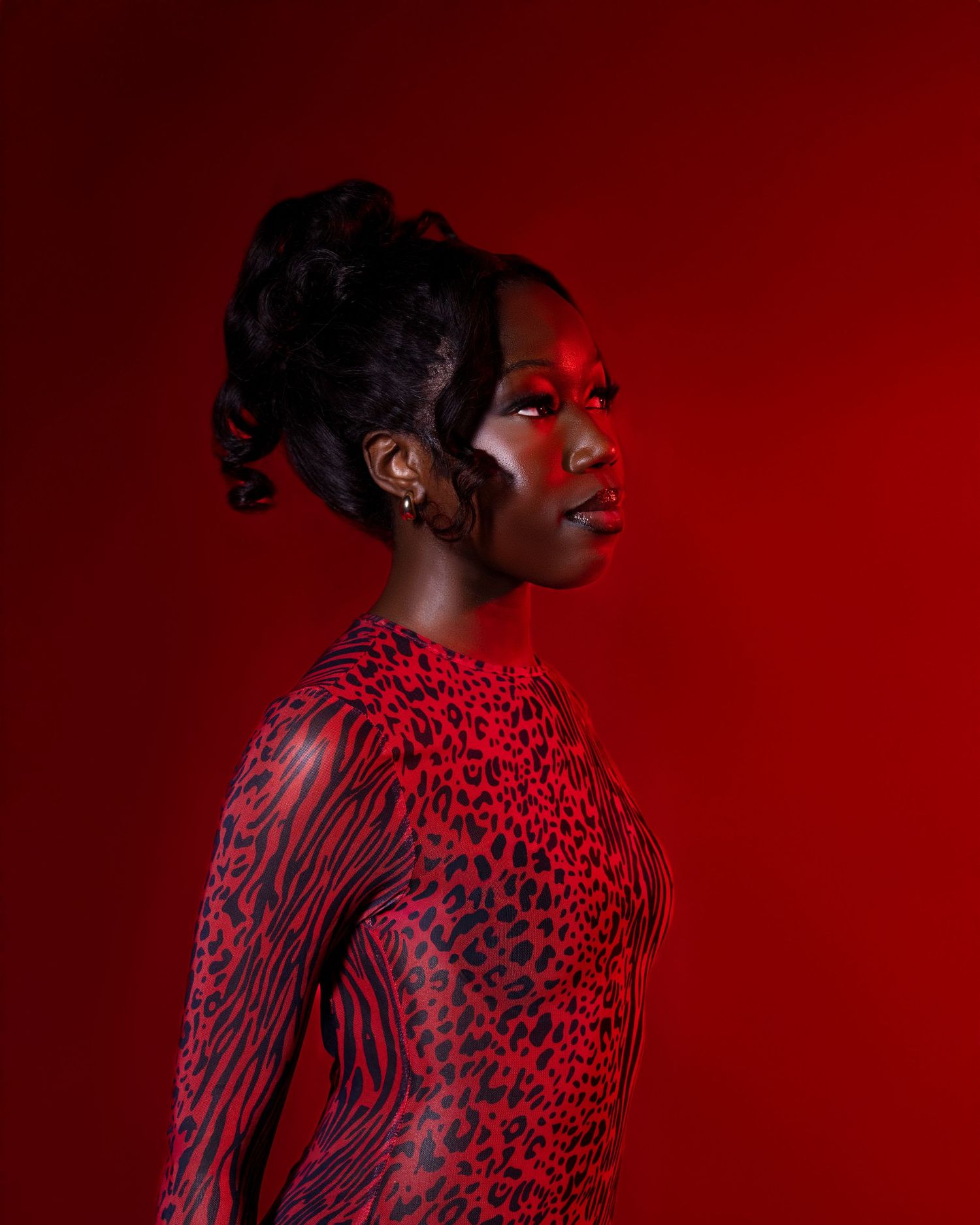 A Black woman in a red leopard-print bodysuit poses in profile against a bold red background with dramatic red lighting.