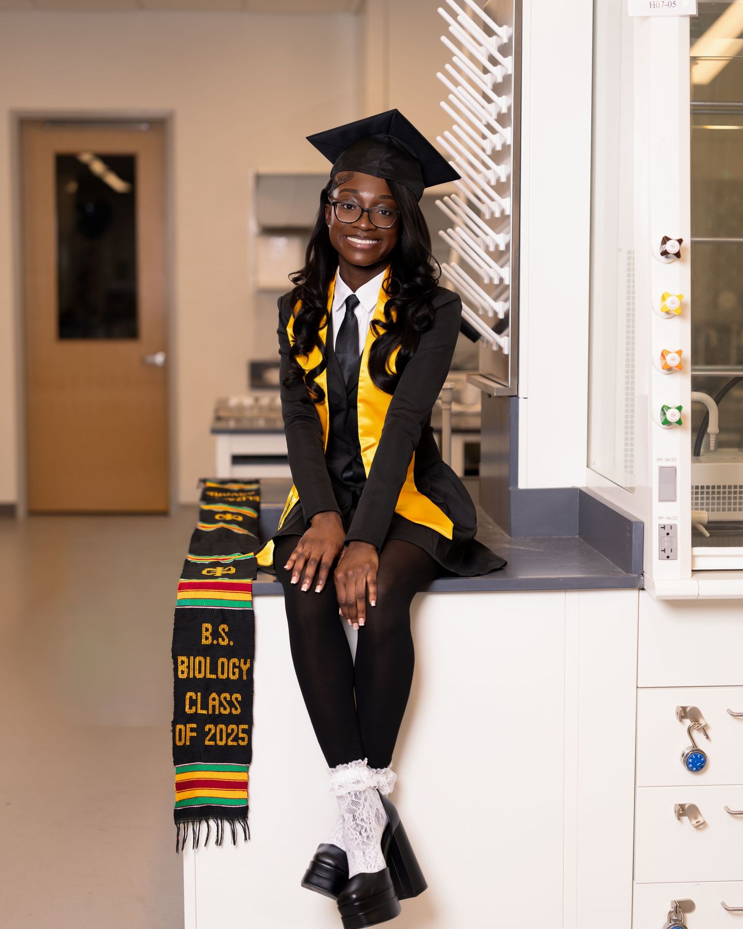 A proud graduate poses in cap and gown with a B.S. Biology Class of 2025 stole in a science laboratory.