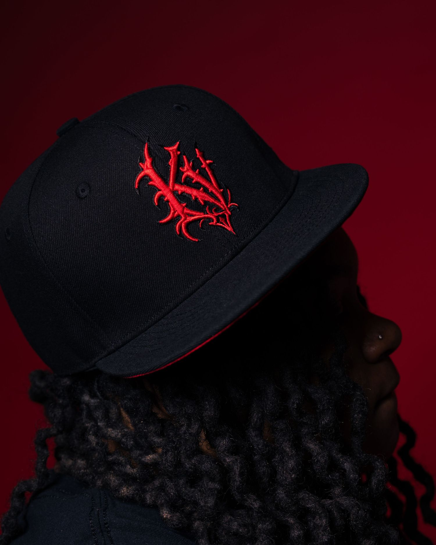 Black fitted cap with red gothic embroidered logo worn by person with dreadlocks against red background.