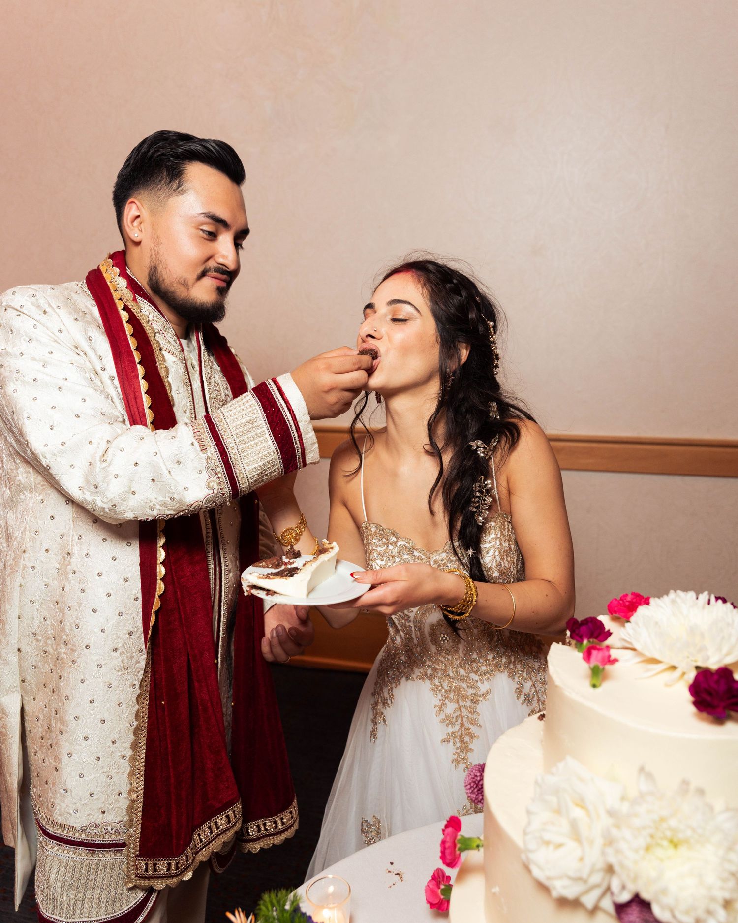 Groom in sherwani feeds wedding cake to bride in gold gown during traditional Indian wedding reception.