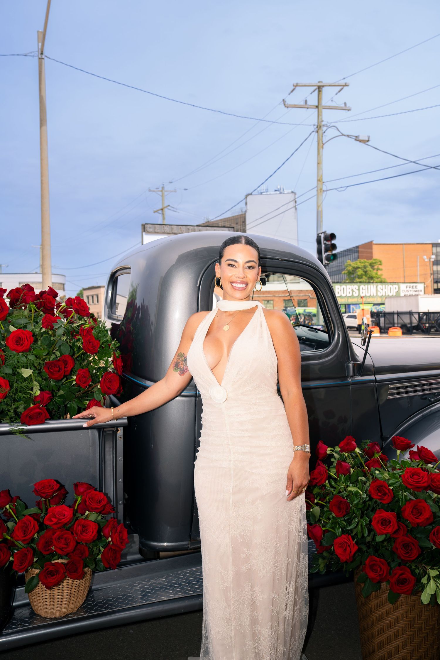 Woman in white halter gown poses by vintage black truck surrounded by red roses on a city street.