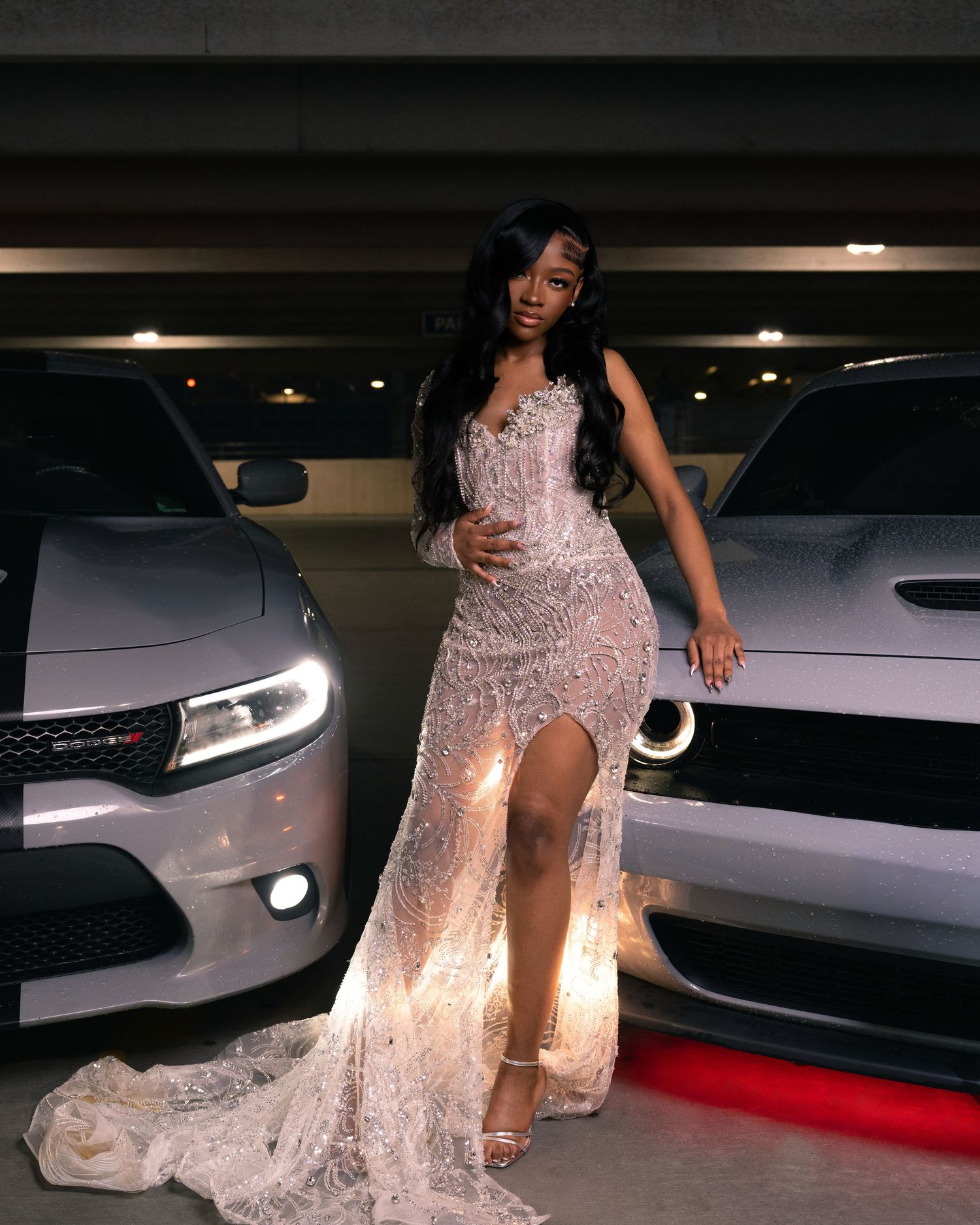 Woman in elegant beaded gown poses between two Dodge muscle cars in a dimly lit parking garage.