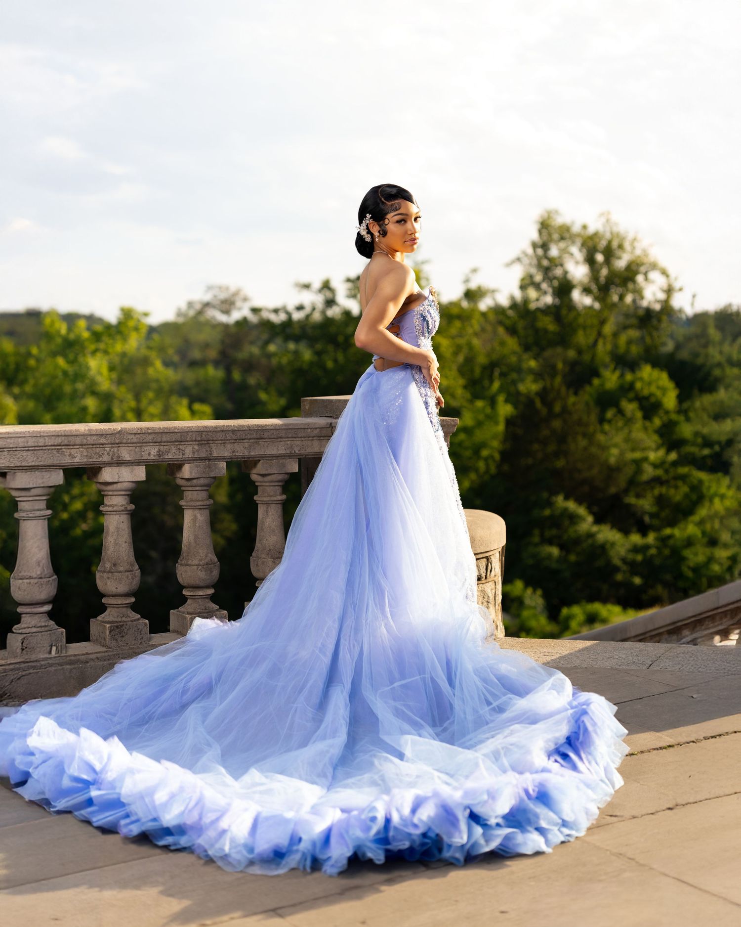 Woman in a stunning lavender tulle ball gown with ruffled train poses on stone balcony with lush greenery backdrop.