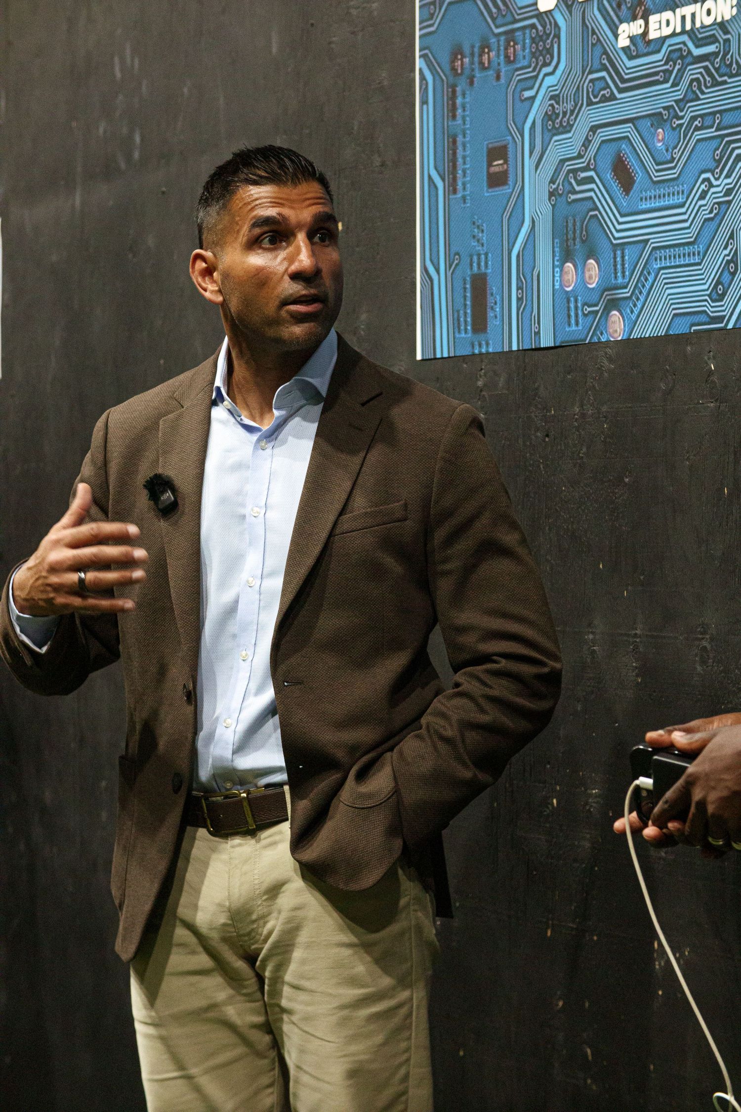 Speaker presenting at a technology conference against a dark wall with circuit board graphics.