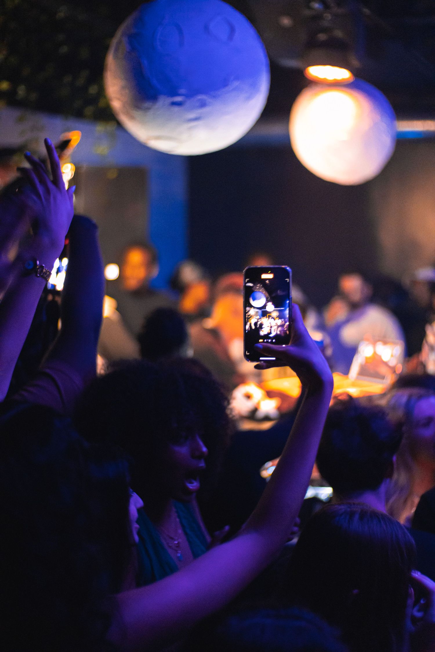 Concert crowd member holds up phone to record while large illuminated spheres hang overhead.