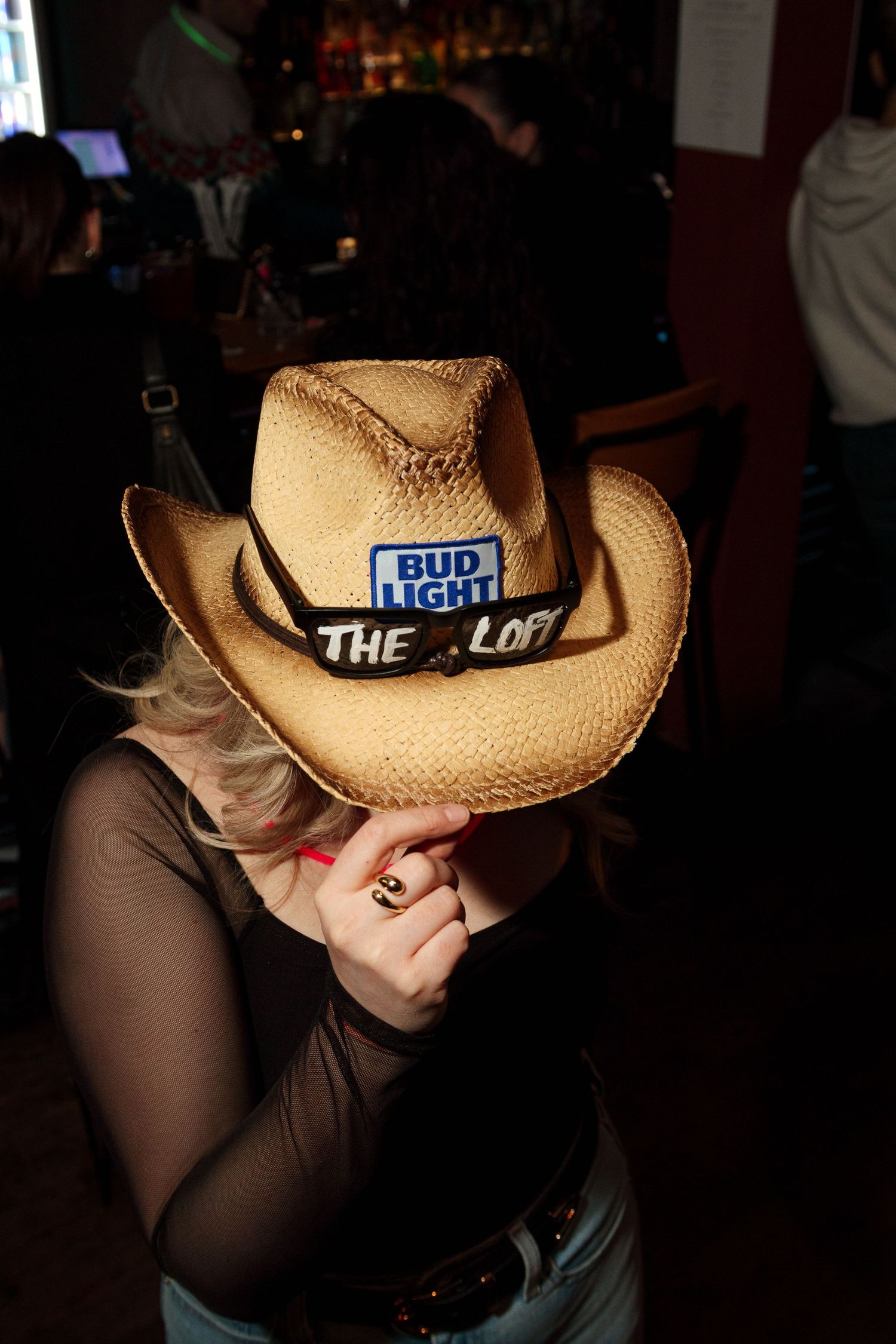 Person wearing a Bud Light promotional cowboy hat in a dimly lit bar setting.