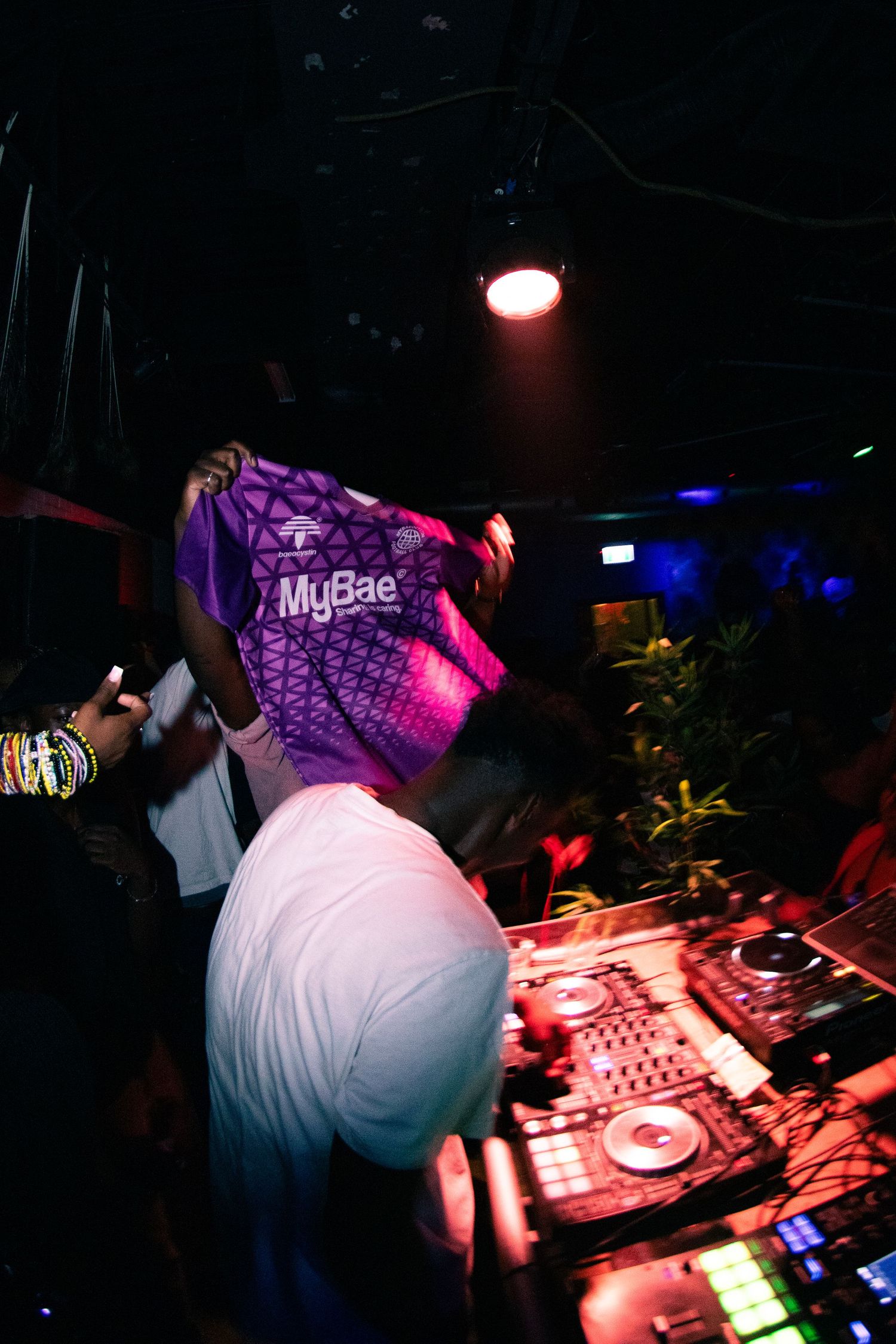 DJ performing with turntables while holding up a purple MyBae shirt in a dark club setting.