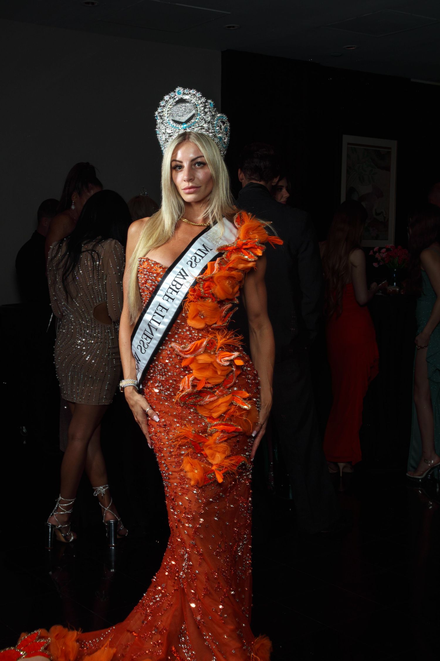 Blonde beauty queen in orange sequin gown with feathers, wearing rhinestone crown and Miss pageant sash at formal event.