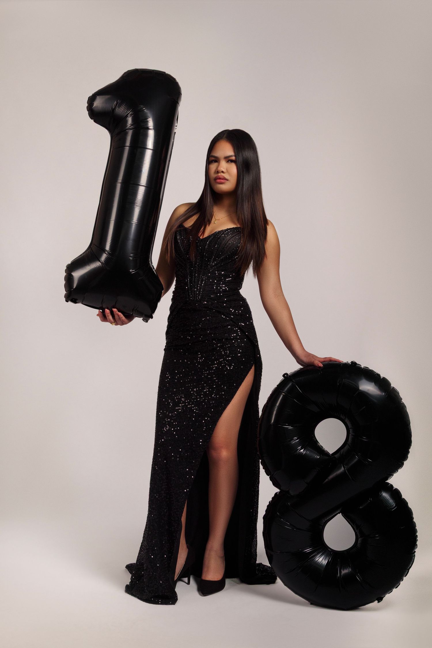 Woman in sparkly black gown posing with large number 18 balloons against light background.